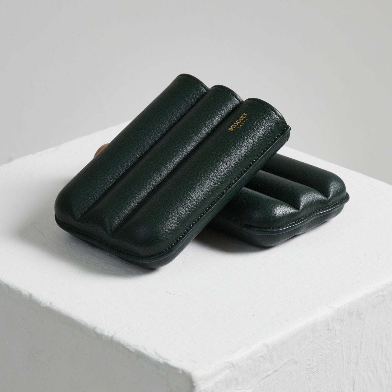 Two green Bosque cigar cases made with French leathers sitting on top of a white cube.