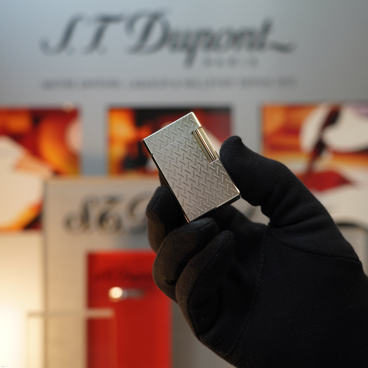 A gloved hand holds a Lucy Lighter Vintage 1990 S.T. Dupont Ligne 1 Large Herringbone Guilloché Heavy Silver Finish lighter, displayed before a blurred S.T. Dupont background—an exquisite collector’s piece.