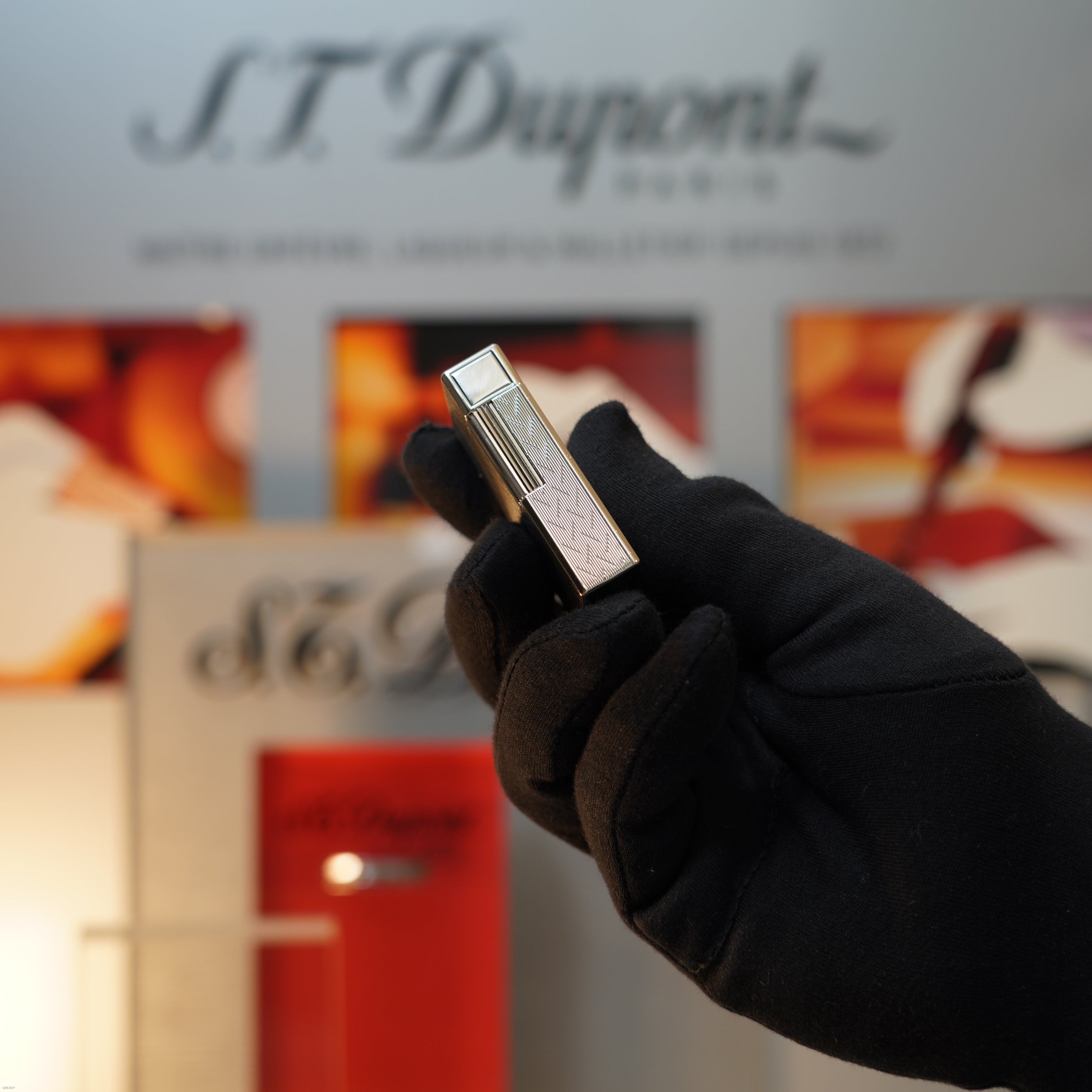 A gloved hand holds a Vintage 1990 S.T. Dupont Ligne 1 Large Herringbone Guilloché Heavy Silver Finish Lighter by Lucy Lighter, with blurred packaging and display in the background.