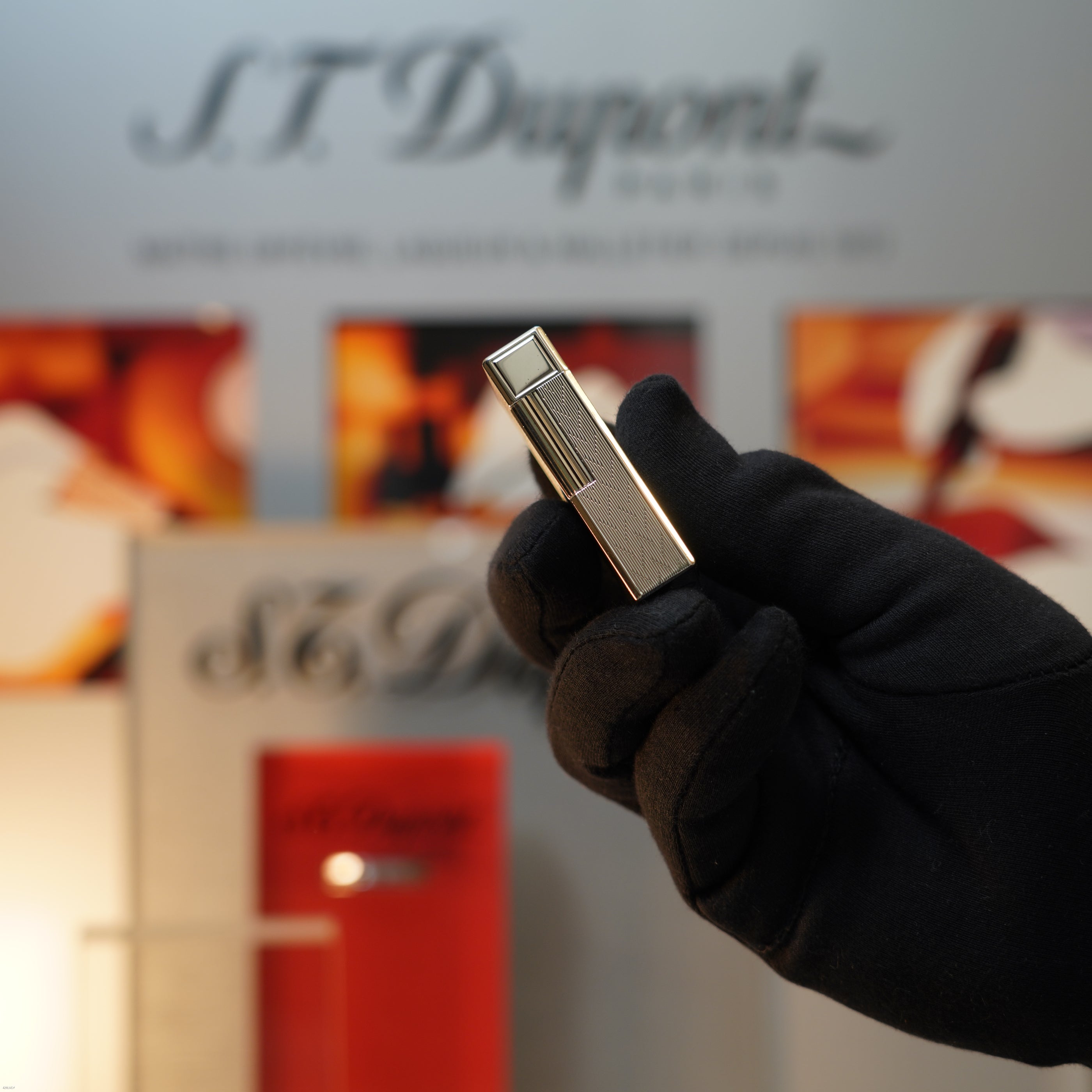 A gloved hand holds a Vintage 1990 S.T. Dupont Ligne 1 Large Herringbone Guilloché Heavy Silver Finish Lighter by Lucy Lighter, with blurred branded packaging and signage in the background.