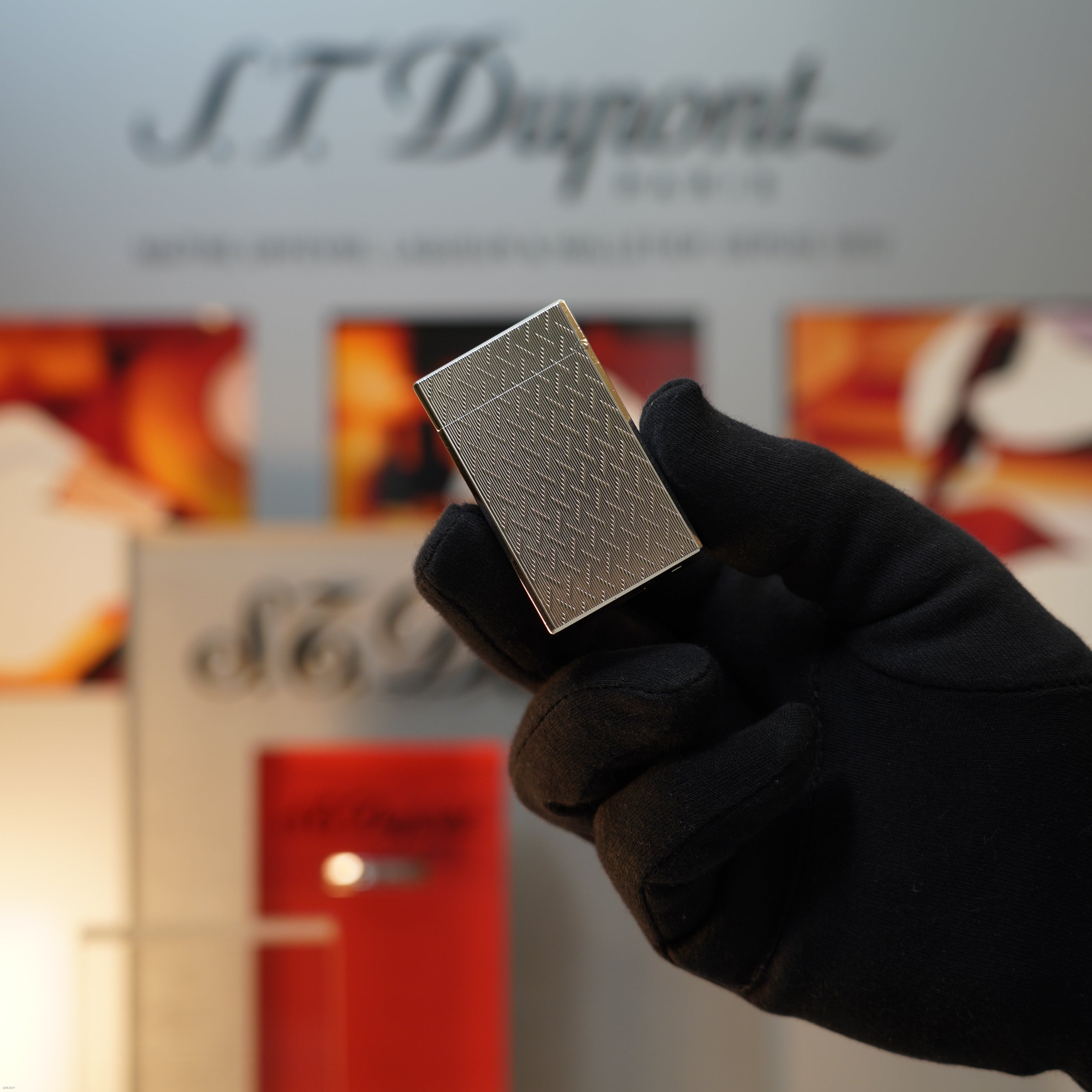 A gloved hand holds a Vintage 1990 S.T. Dupont Ligne 1 Large Herringbone Guilloché Heavy Silver Finish Lighter from Lucy Lighter, with iconic branding visible in the blurred background.