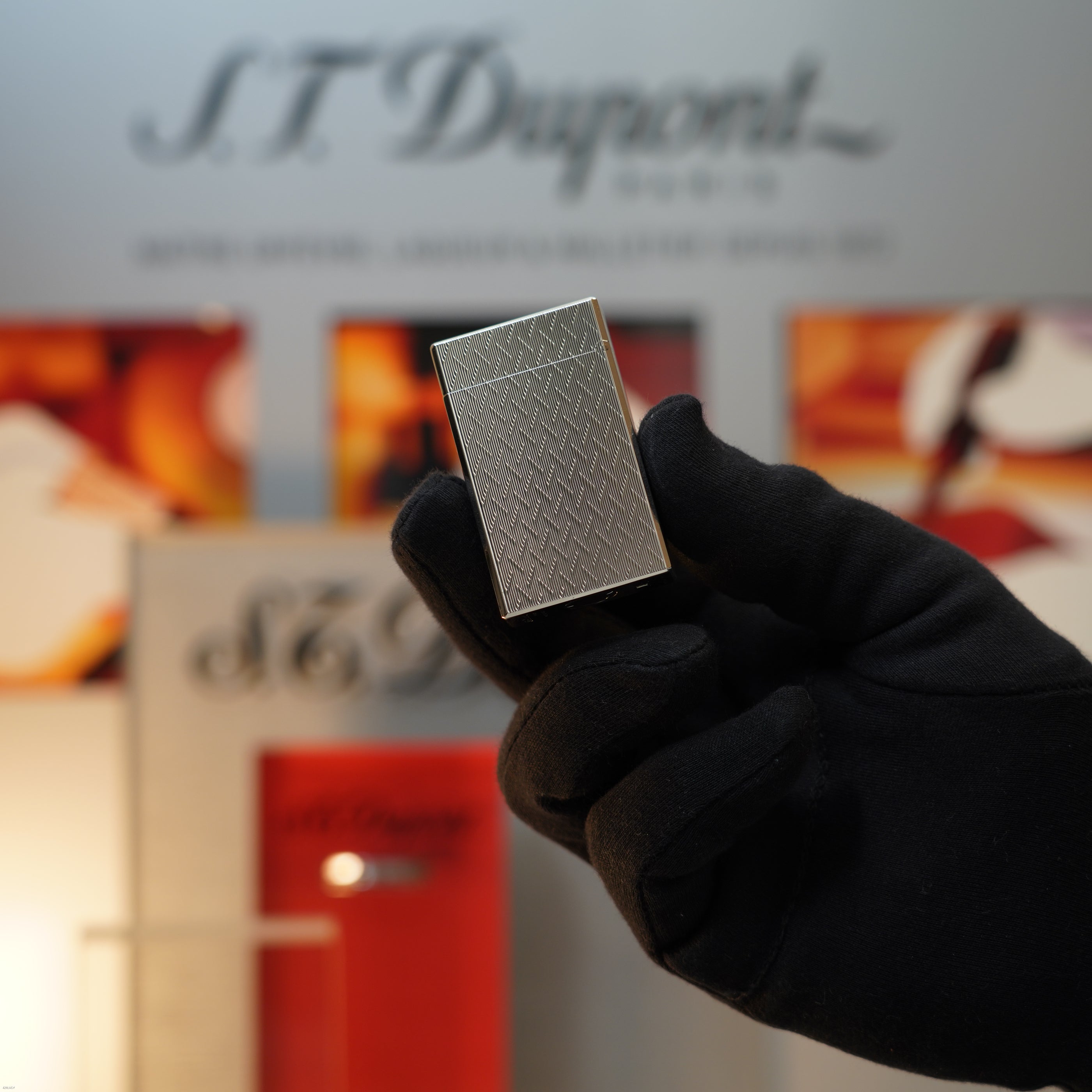 A gloved hand holds a Vintage 1990 S.T. Dupont Ligne 1 Large Herringbone Guilloché Heavy Silver Finish Lighter by Lucy Lighter, its textured pattern shown against a blurred branded backdrop.