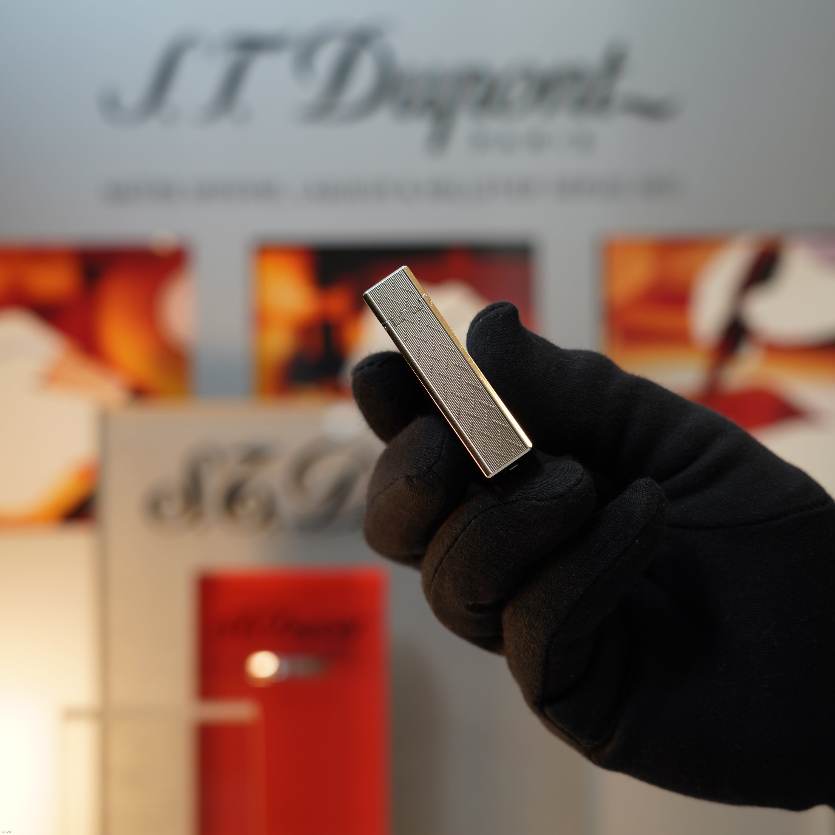 A gloved hand holds a Vintage 1990 Lucy Lighter S.T. Dupont Ligne 1 Large Herringbone Guilloché Heavy Silver Finish lighter against a blurred background with the S.T. Dupont logo and product display.