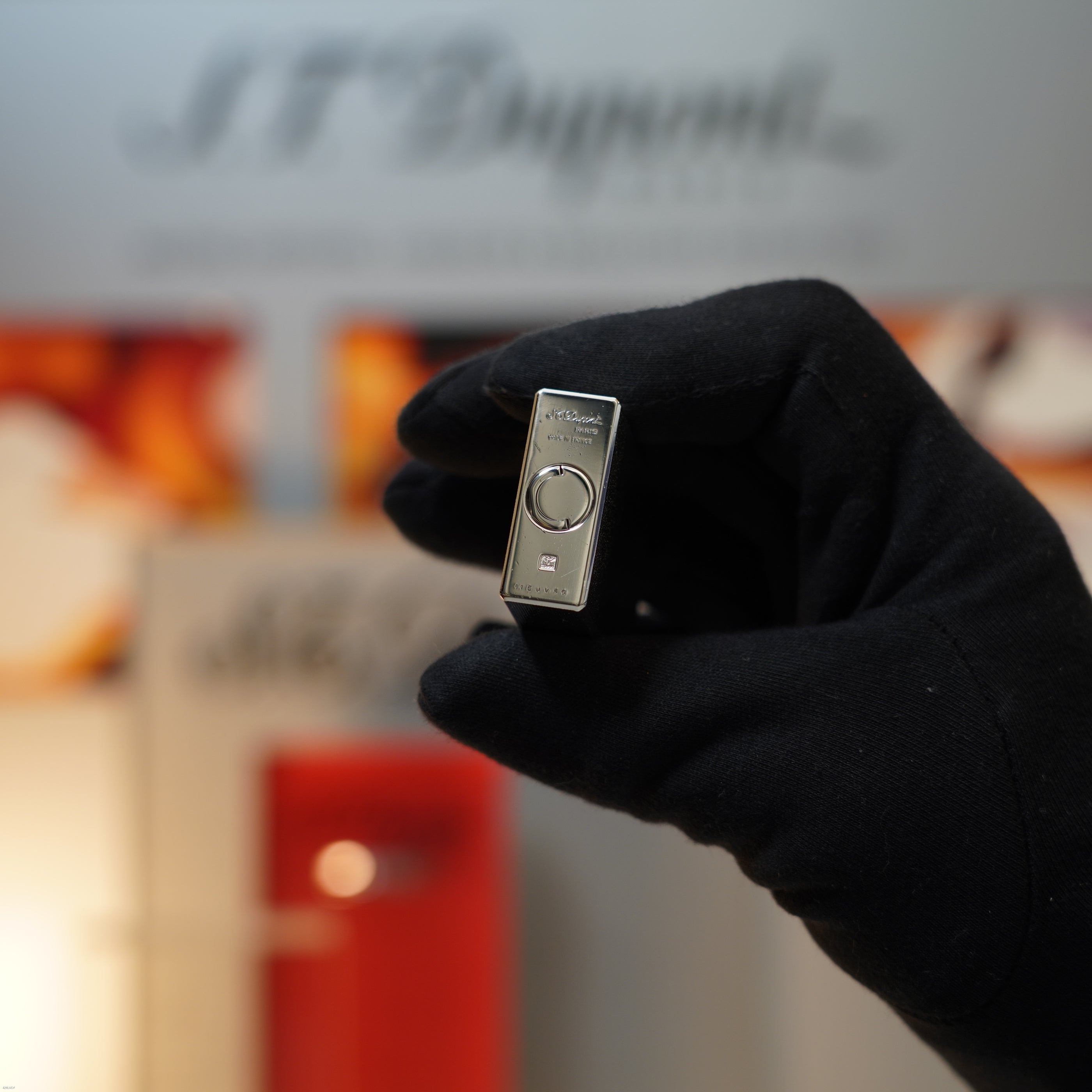 A gloved hand holds a Vintage 1990 Lucy Lighter Ligne 1 Large Herringbone Guilloché heavy silver finish lighter with a circular button, against a blurred background—a prized collector’s piece.