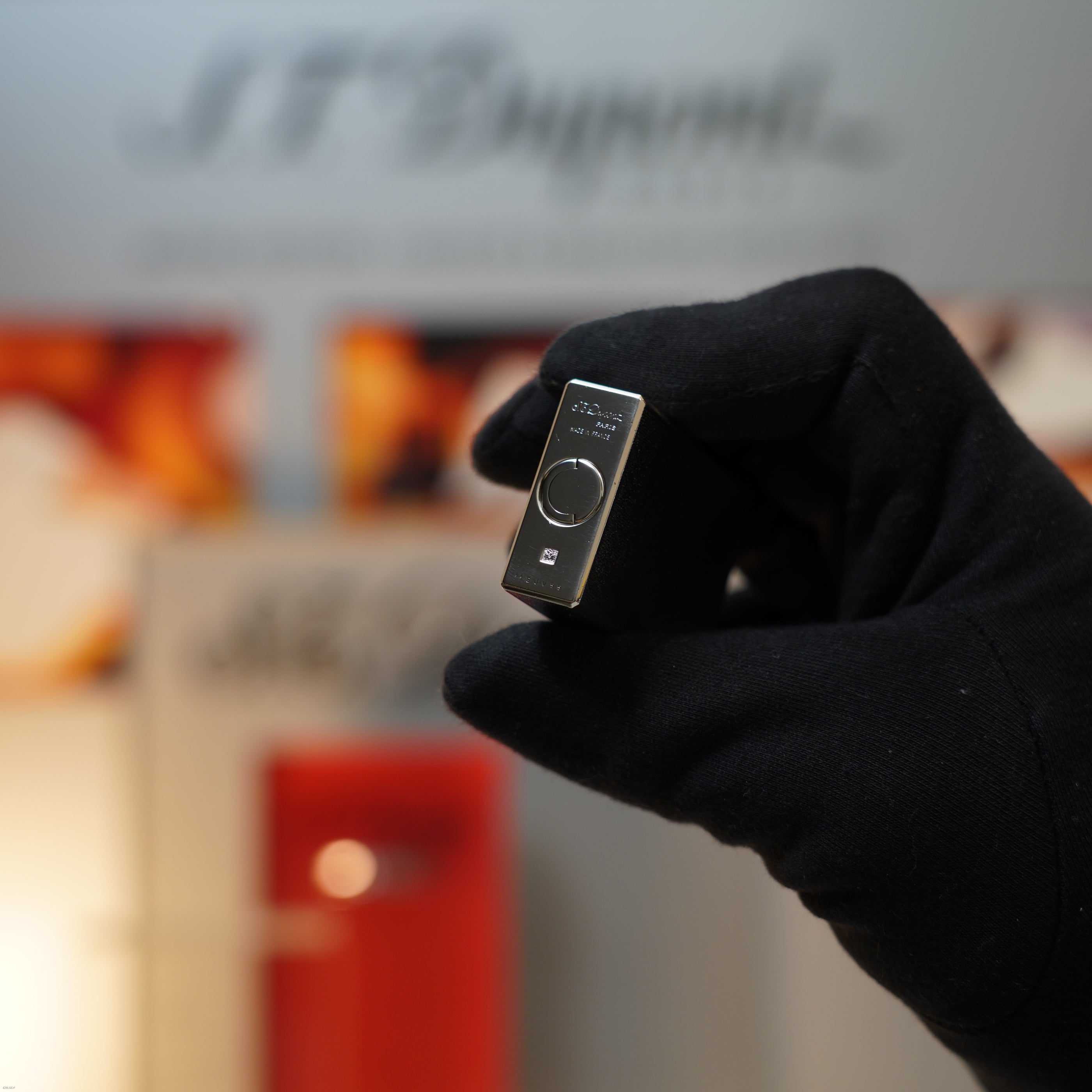 A gloved hand holds a Vintage 1990 S.T. Dupont Ligne 1 Large Herringbone Guilloché Heavy Silver Finish Lighter by Lucy Lighter, shown against a blurred backdrop featuring the words "ST Dupont.
