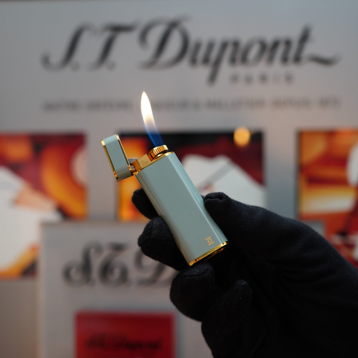 A gloved hand holds a lit Cartier luxury lighter, the Vintage 1980 Oval with a 24k gold finish and gris gray lacquer from the Le Must series, elegantly showcasing its flame.