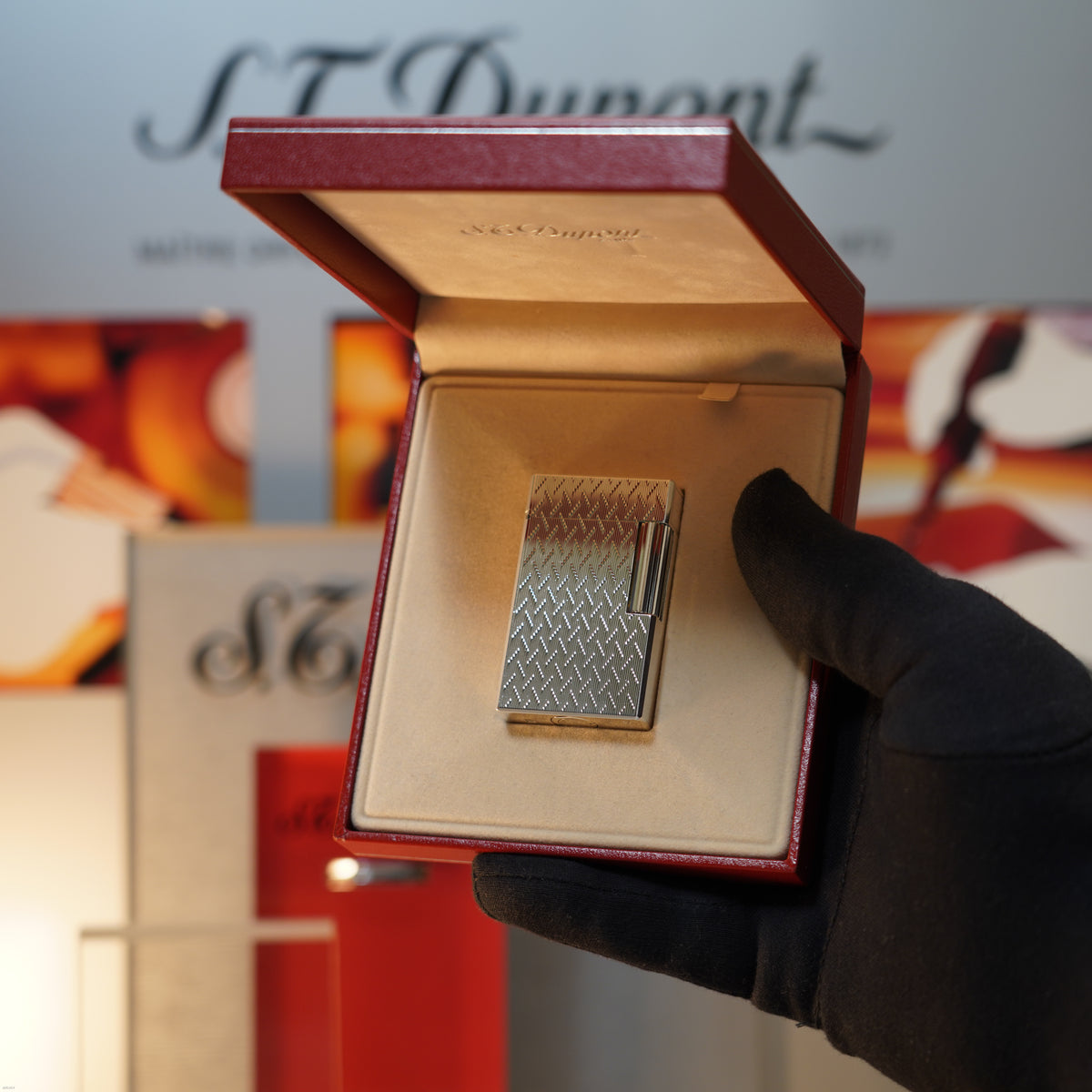 A gloved hand holds an open red box showing a Vintage 1990 Lucy Lighter S.T. Dupont Ligne 1 Large Herringbone Guilloché Heavy Silver Finish Lighter, with a blurred background featuring the S.T. Dupont logo.