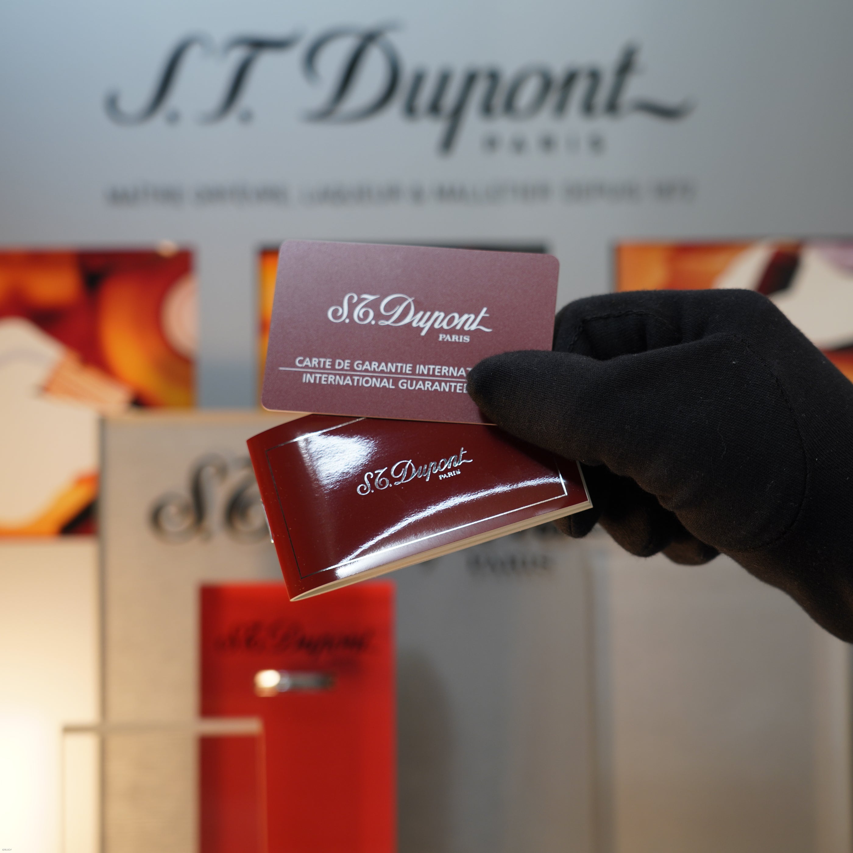 A gloved hand holds two Lucy Lighter cards in front of packaging and products, including a Vintage 1990 Lucy Lighter Ligne 1 Large Herringbone Guilloché Heavy Silver Finish Lighter, on display.