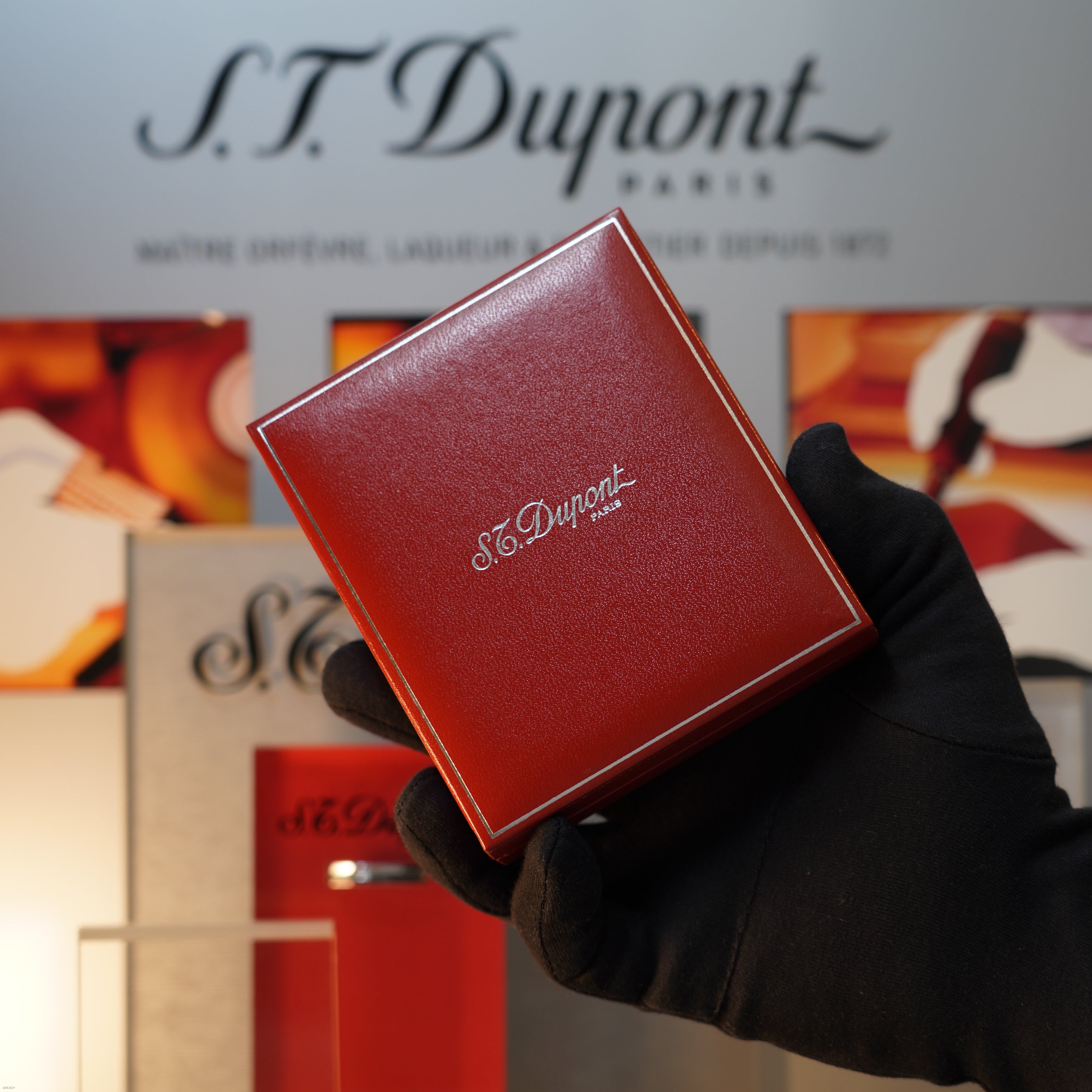 A gloved hand holds a red Lucy Lighter box, possibly containing a Vintage 1990 S.T. Dupont Ligne 1 Large Herringbone Guilloché Heavy Silver Finish Lighter, in front of a display with the S.T. Dupont logo and branded decor.