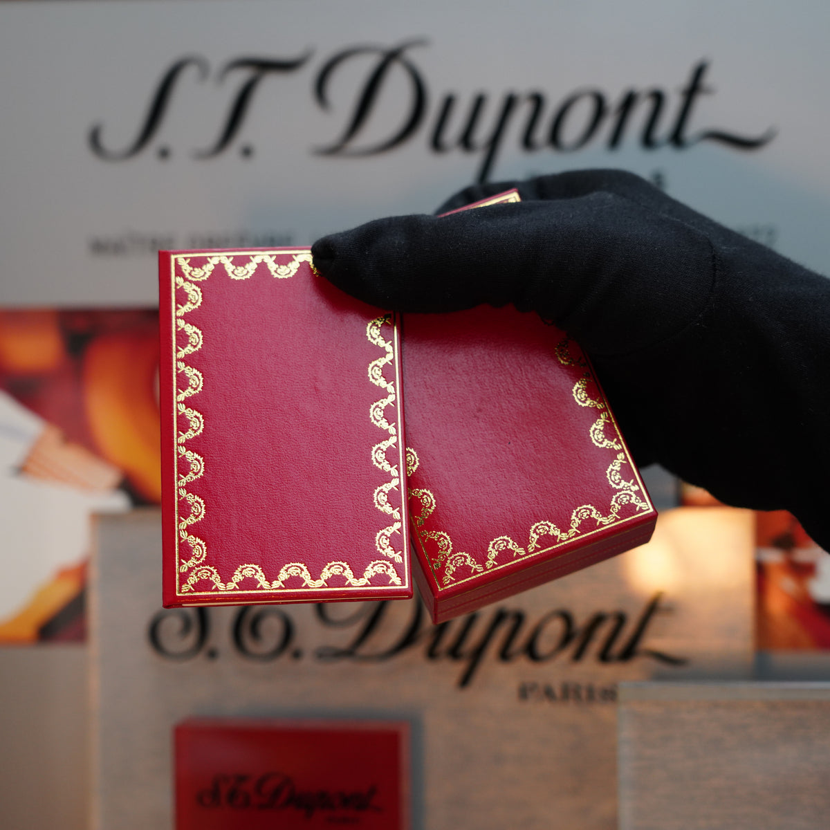 A gloved hand holds an open red box with gold designs, presenting a Vintage 1980 Cartier Oval 24k Gold Finish gris gray Lacquer Le Must Lighter in front of blurred text featuring "Cartier.