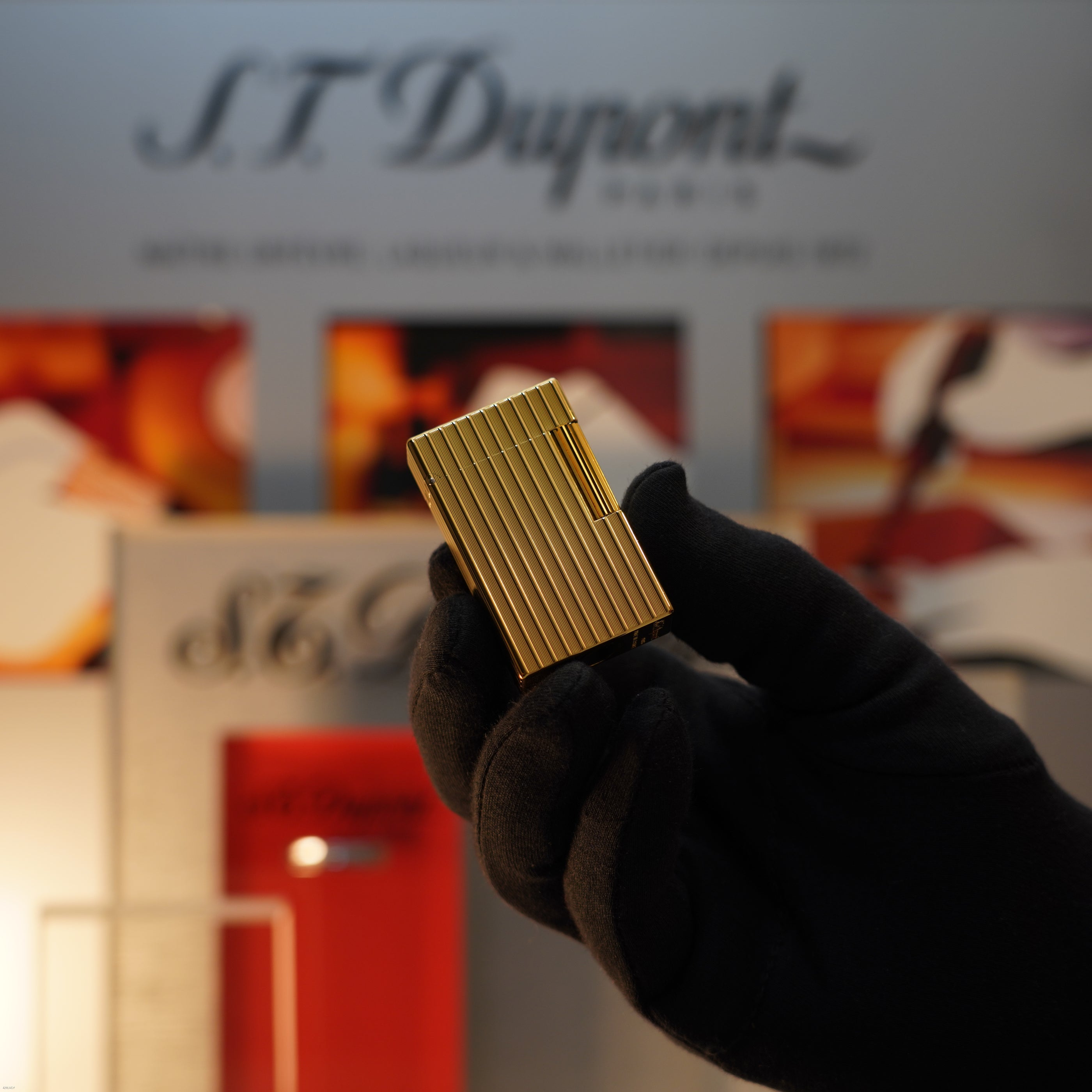 A gloved hand holds a Vintage November 1987 S.T. Dupont Large Ligne 1 Heavy rare pattern 18k Gold Finish Lighter by Lucy Lighter in front of a blurred S.T. Dupont display.