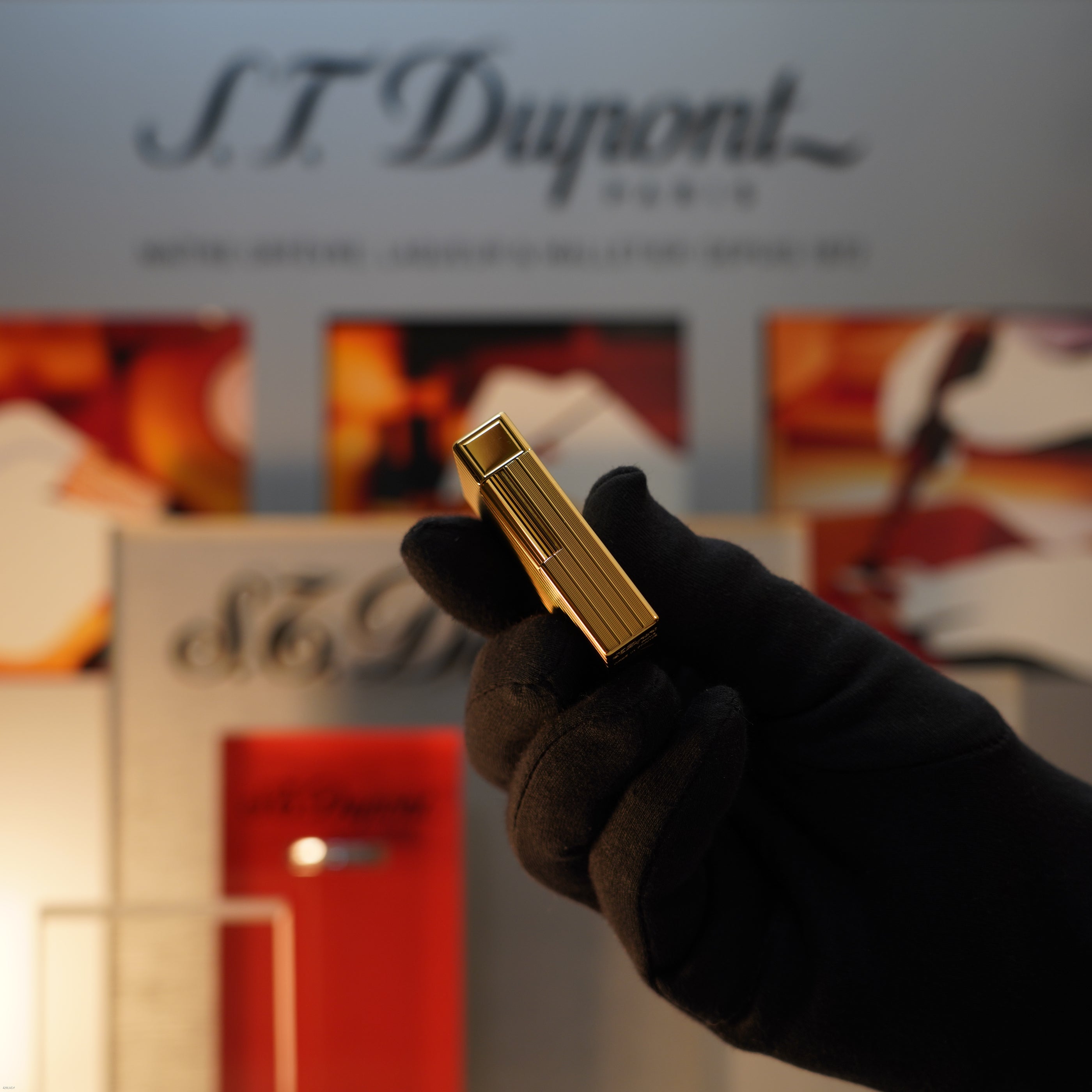 A gloved hand holds a Vintage November 1987 Lucy Lighter Large Ligne 1 with a rare pattern and 18k gold finish, in front of a blurred S.T. Dupont display.