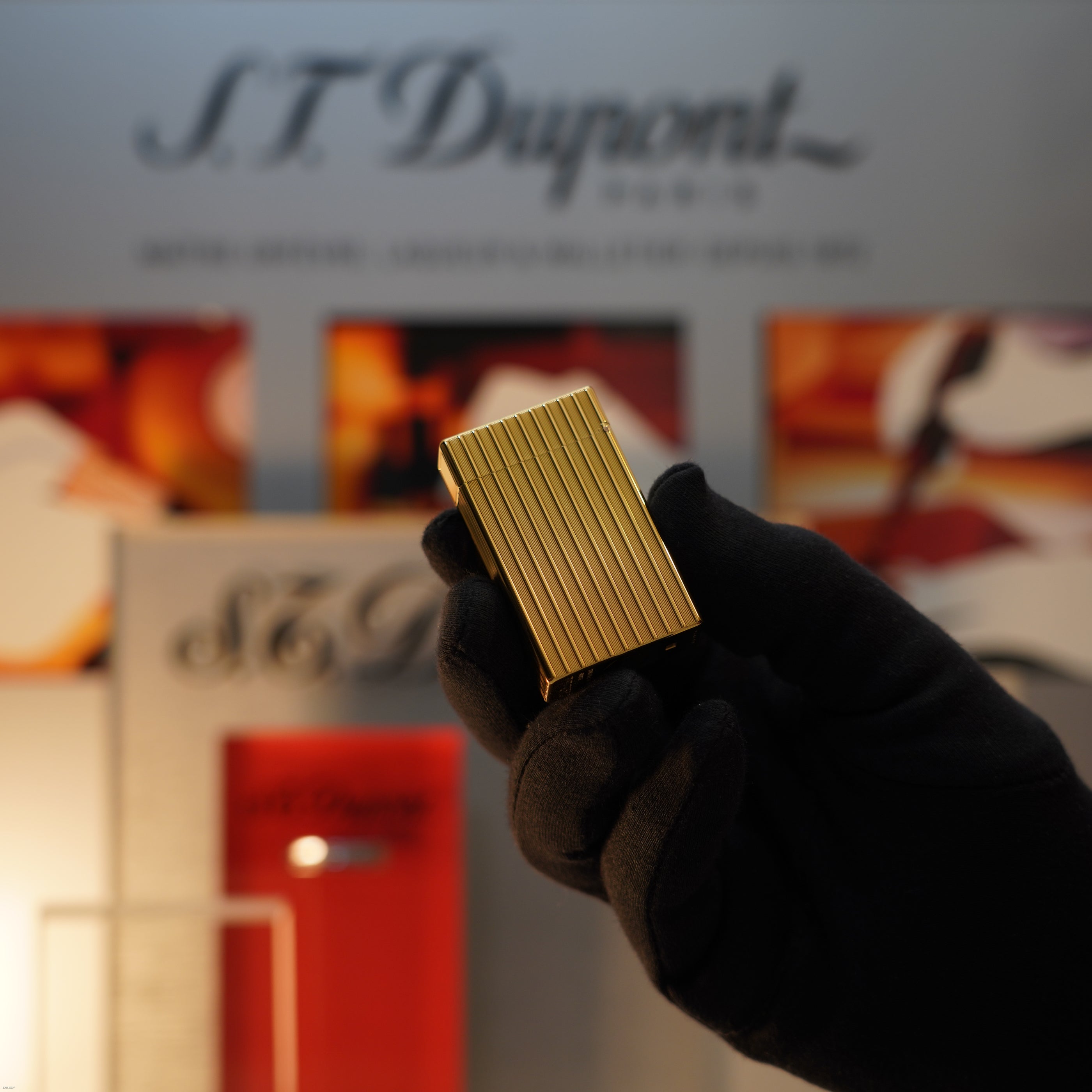 A gloved hand holds a Vintage November 1987 Lucy Lighter Large Ligne 1 with a rare heavy pattern and 18k gold finish, set against a blurred S.T. Dupont display in the background.