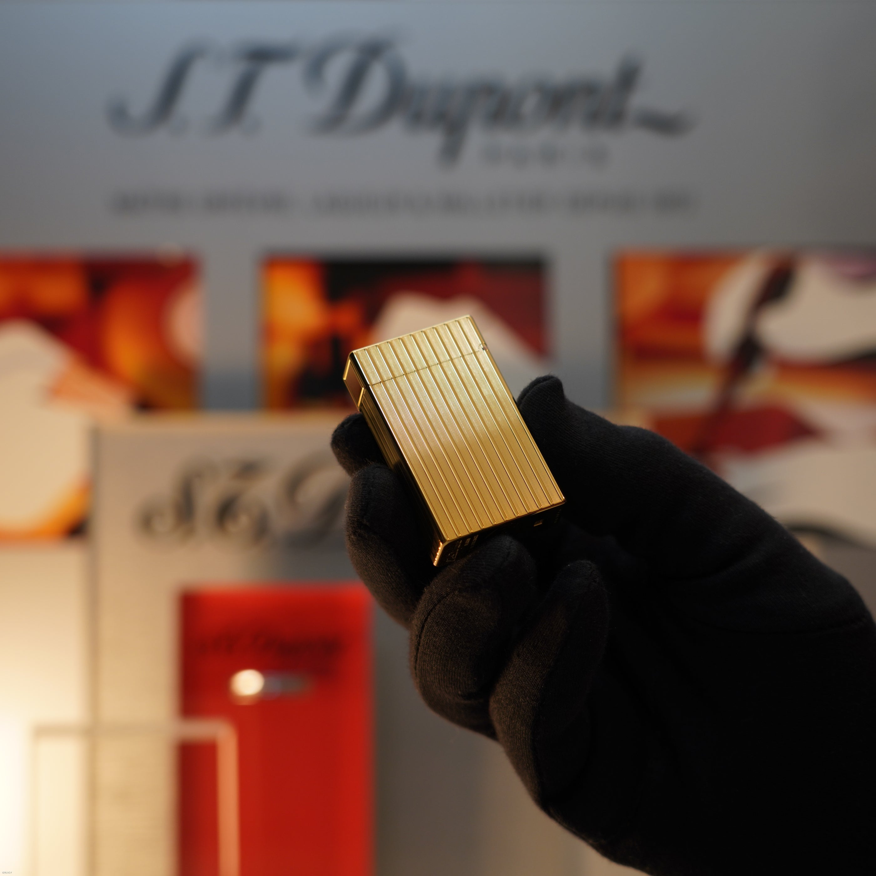 A gloved hand holds a Vintage November 1987 S.T. Dupont Large Ligne 1 Heavy rare pattern 18k Gold Finish Lighter by Lucy Lighter, shown against a blurred S.T. Dupont Paris display background.