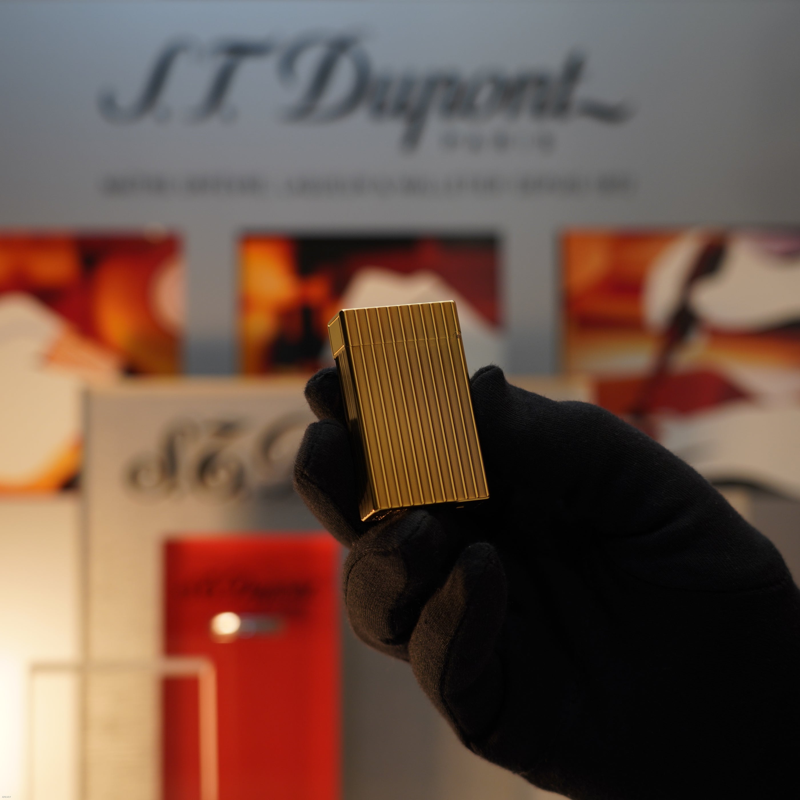 A gloved hand holds a Lucy Lighter Vintage November 1987 S.T. Dupont Large Ligne 1 heavy rare pattern lighter with an 18k gold finish, set against a blurred S.T. Dupont Paris display background.