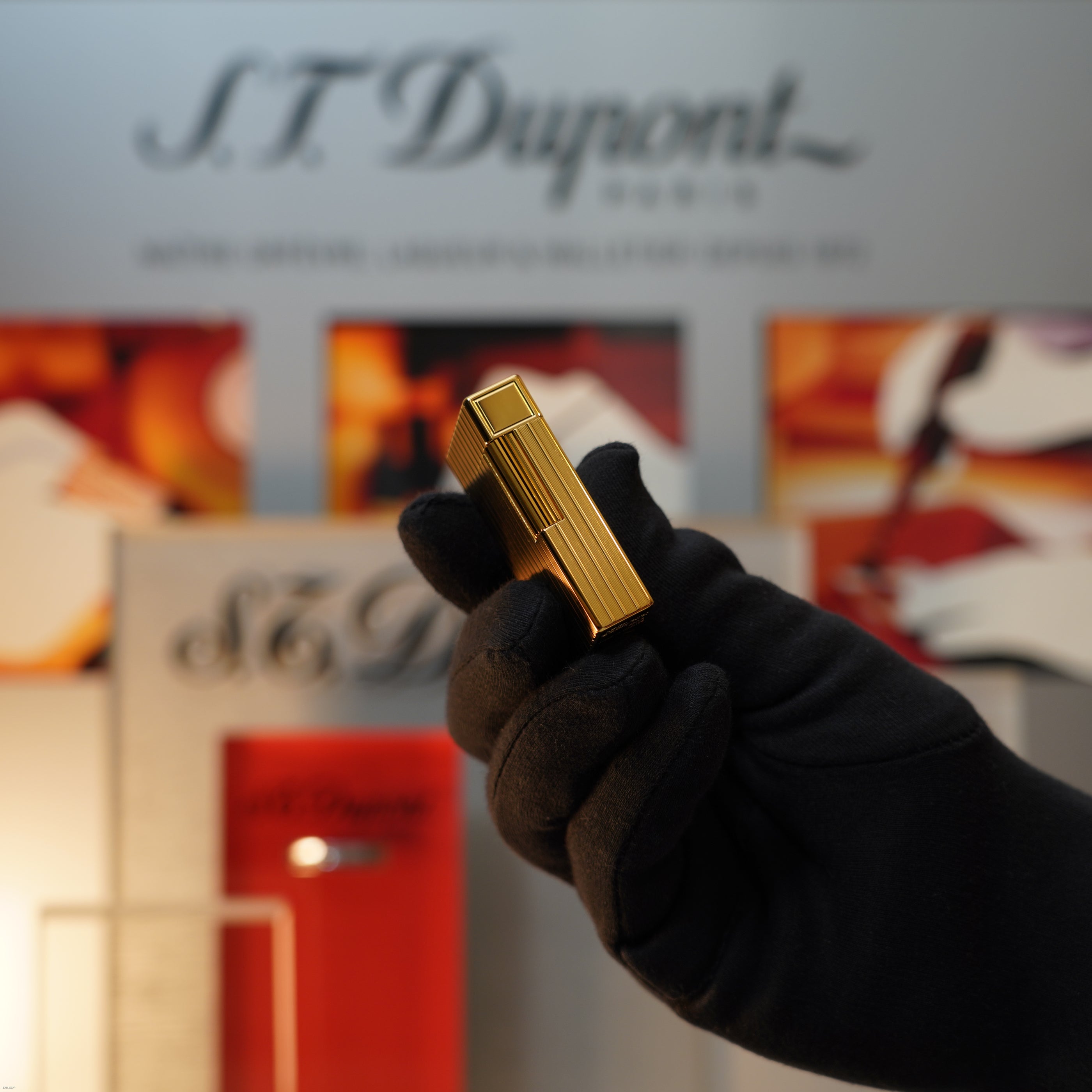 A gloved hand holds a Lucy Lighter Vintage November 1987 S.T. Dupont Large Ligne 1 Heavy rare pattern 18k Gold Finish Lighter before a blurred S.T. Dupont Paris display.
