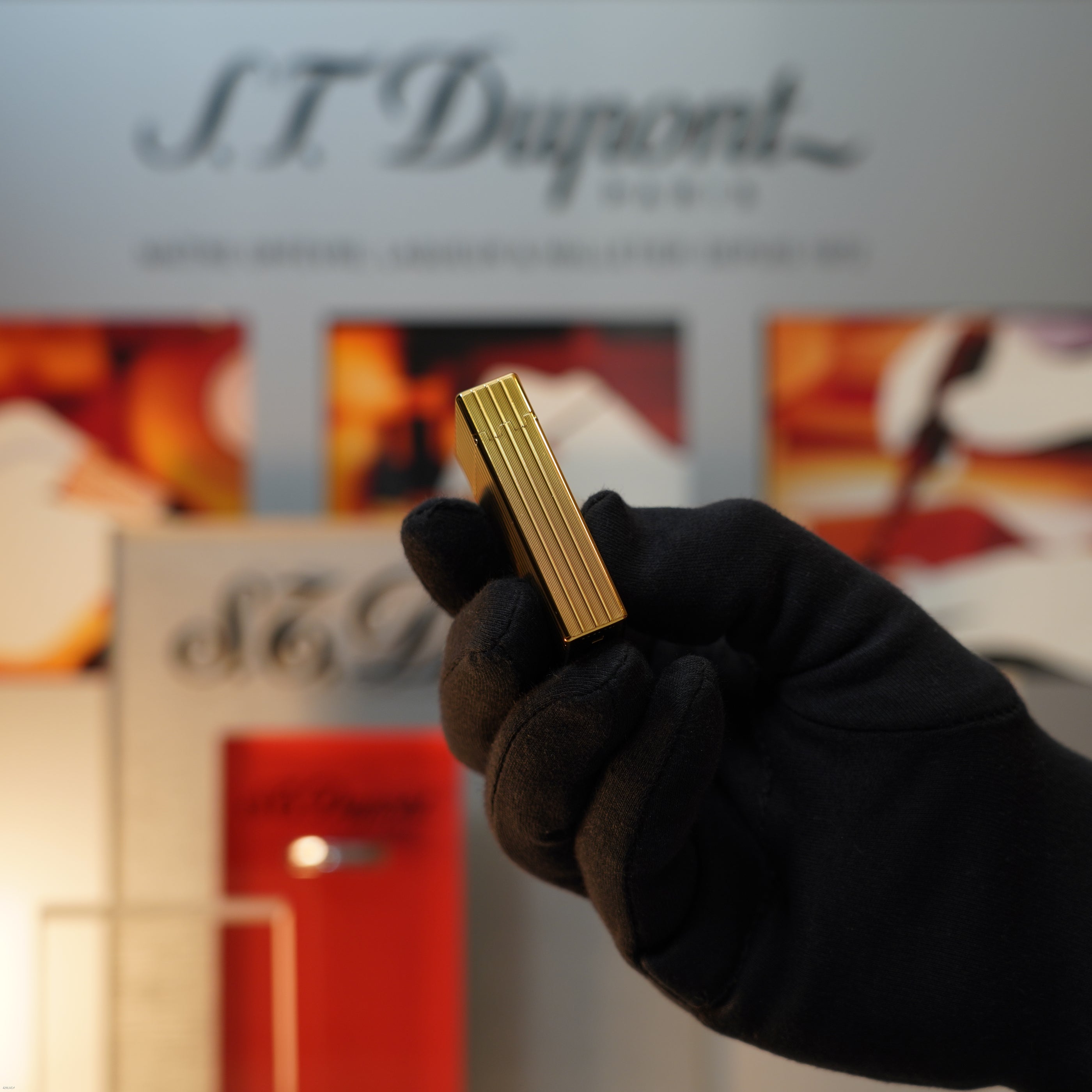 A gloved hand holds a Vintage November 1987 S.T. Dupont Large Ligne 1 Heavy rare pattern 18k Gold Finish Lighter by Lucy Lighter, with a blurred display of S.T. Dupont products in the background.