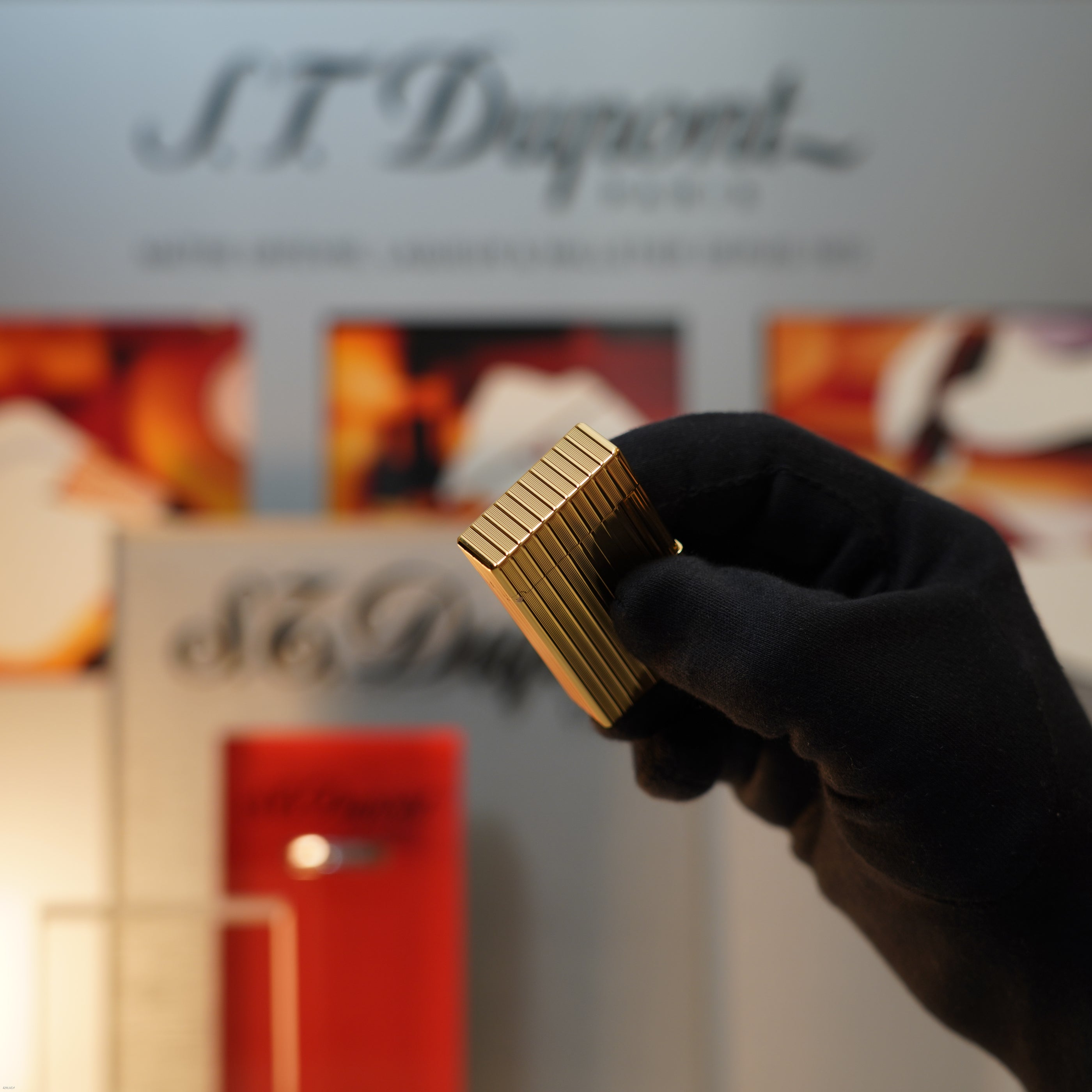 A gloved hand holds a Vintage November 1987 Lucy Lighter Large Ligne 1 rare pattern lighter with an 18k gold finish in front of a blurred S.T. Dupont display.