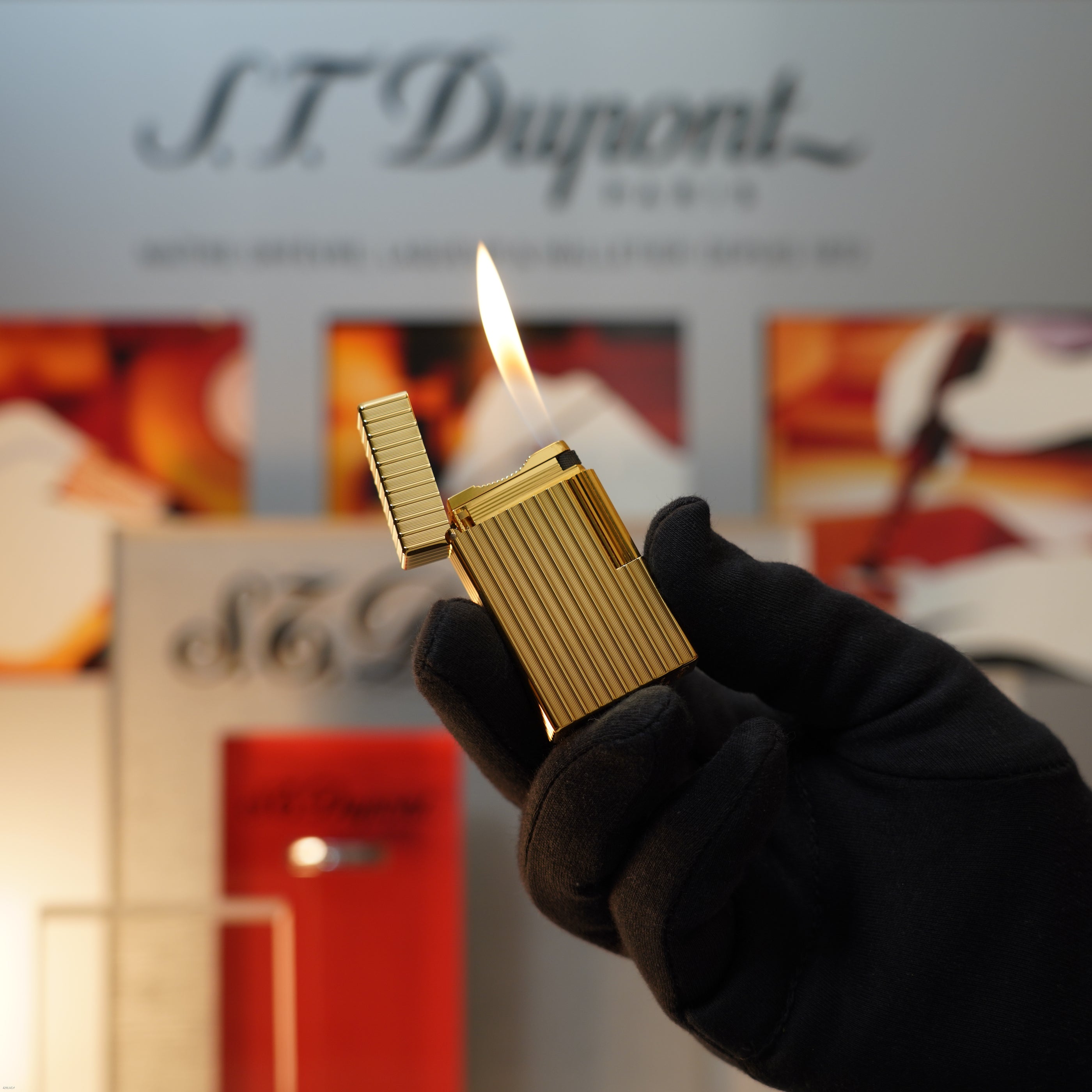 A gloved hand holds a lit Lucy Lighter Vintage November 1987 S.T. Dupont Large Ligne 1 Heavy rare pattern 18k Gold Finish Lighter, with branded packaging softly blurred in the background.