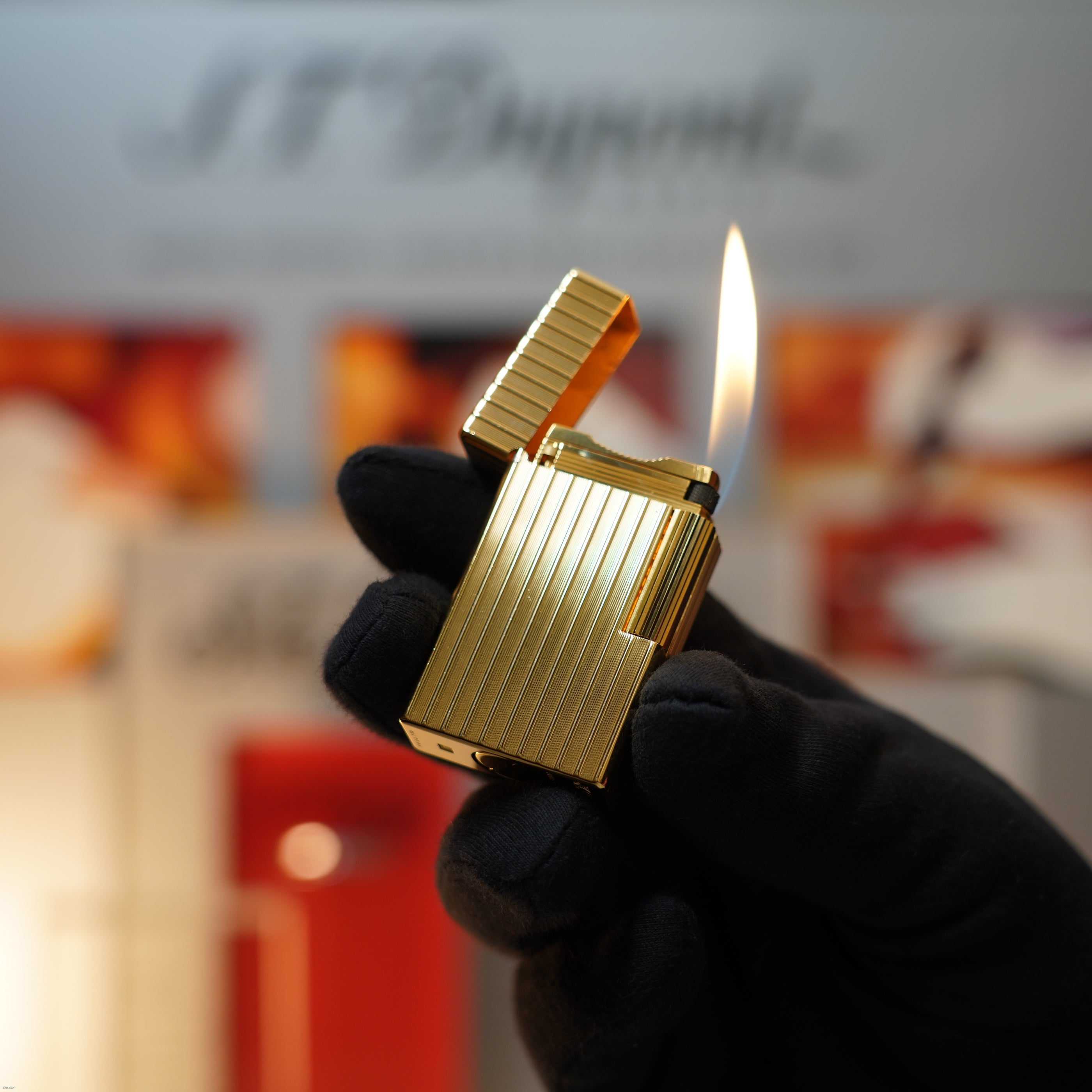 A gloved hand holds a Lucy Lighter Vintage November 1987 S.T. Dupont Large Ligne 1 Heavy rare pattern lighter, featuring an 18k gold finish and an open ridged lid with a bright flame against a blurred background.