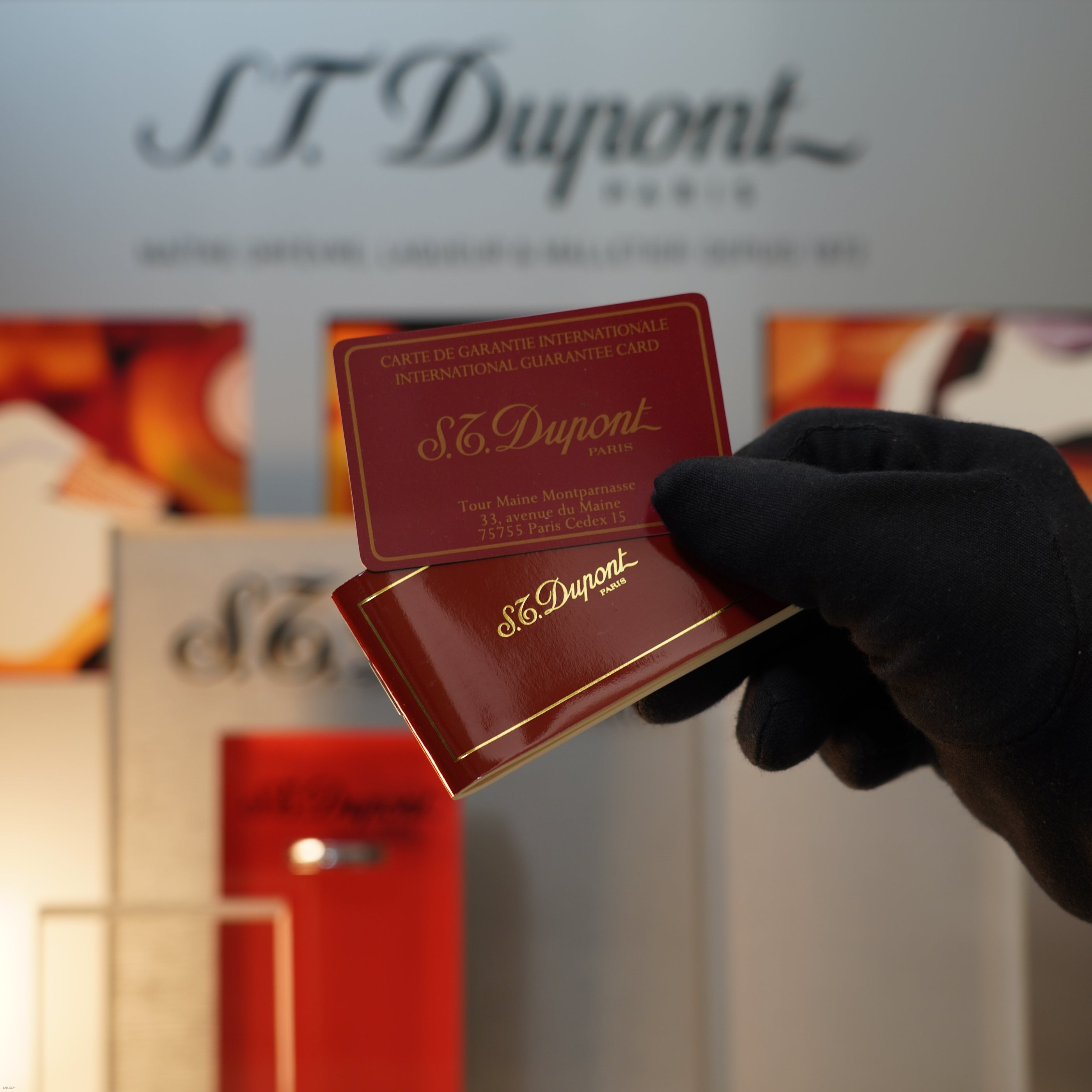 A gloved hand holds a Vintage November 1987 S.T. Dupont Large Ligne 1 Heavy rare pattern 18k Gold Finish Lighter by Lucy Lighter, along with an international guarantee card, in front of a branded display.