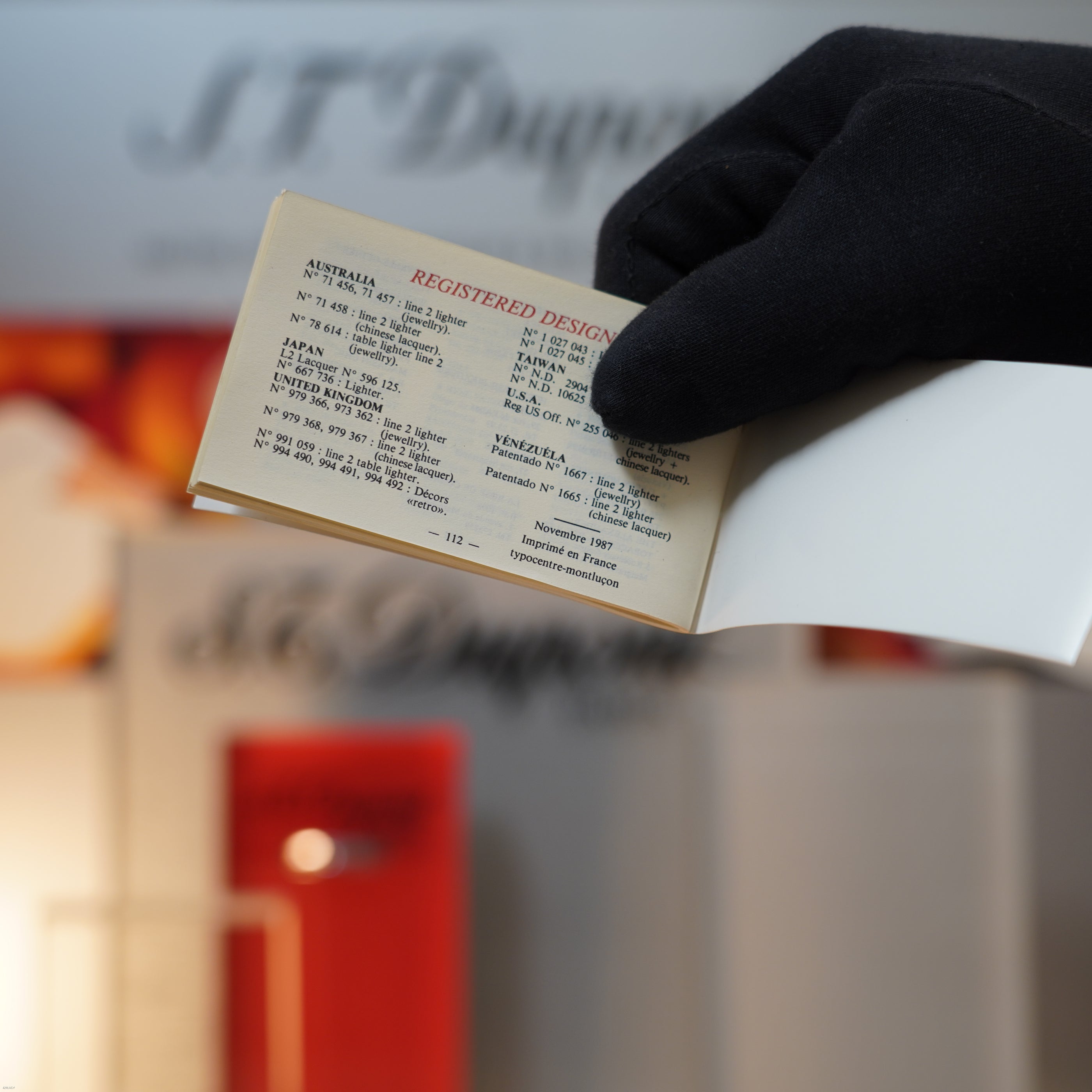 A gloved hand holds an open book showing registered design info, with a blurred "Lucy Lighter" sign suggesting the elegance of the Vintage November 1987 S.T. Dupont Large Ligne 1 Heavy rare pattern 18k Gold Finish Lighter.