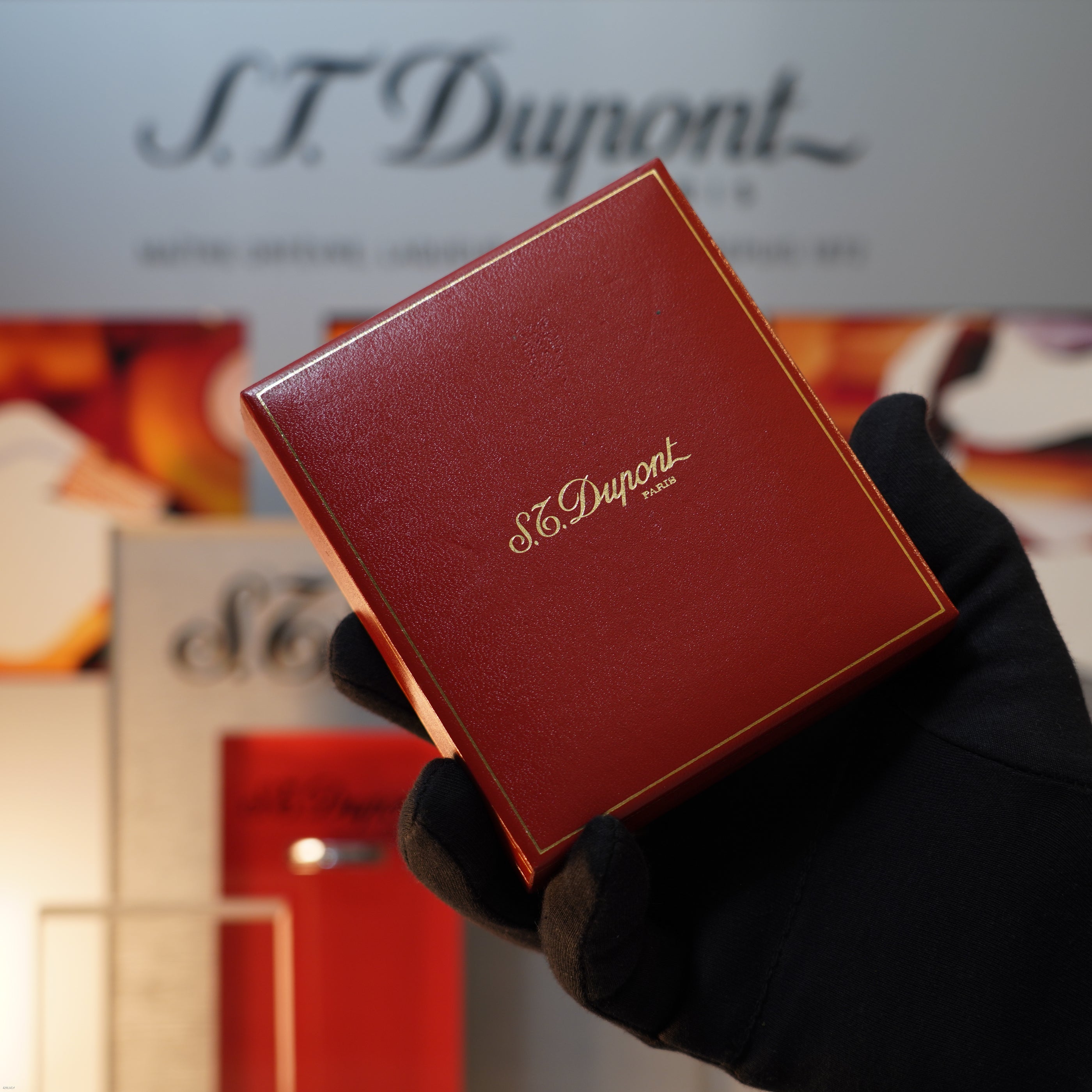 A gloved hand holds a small red box with gold lettering from Lucy Lighter, possibly containing a Vintage November 1987 S.T. Dupont Large Ligne 1 Heavy rare pattern 18k Gold Finish Lighter, set against a blurred branded background.