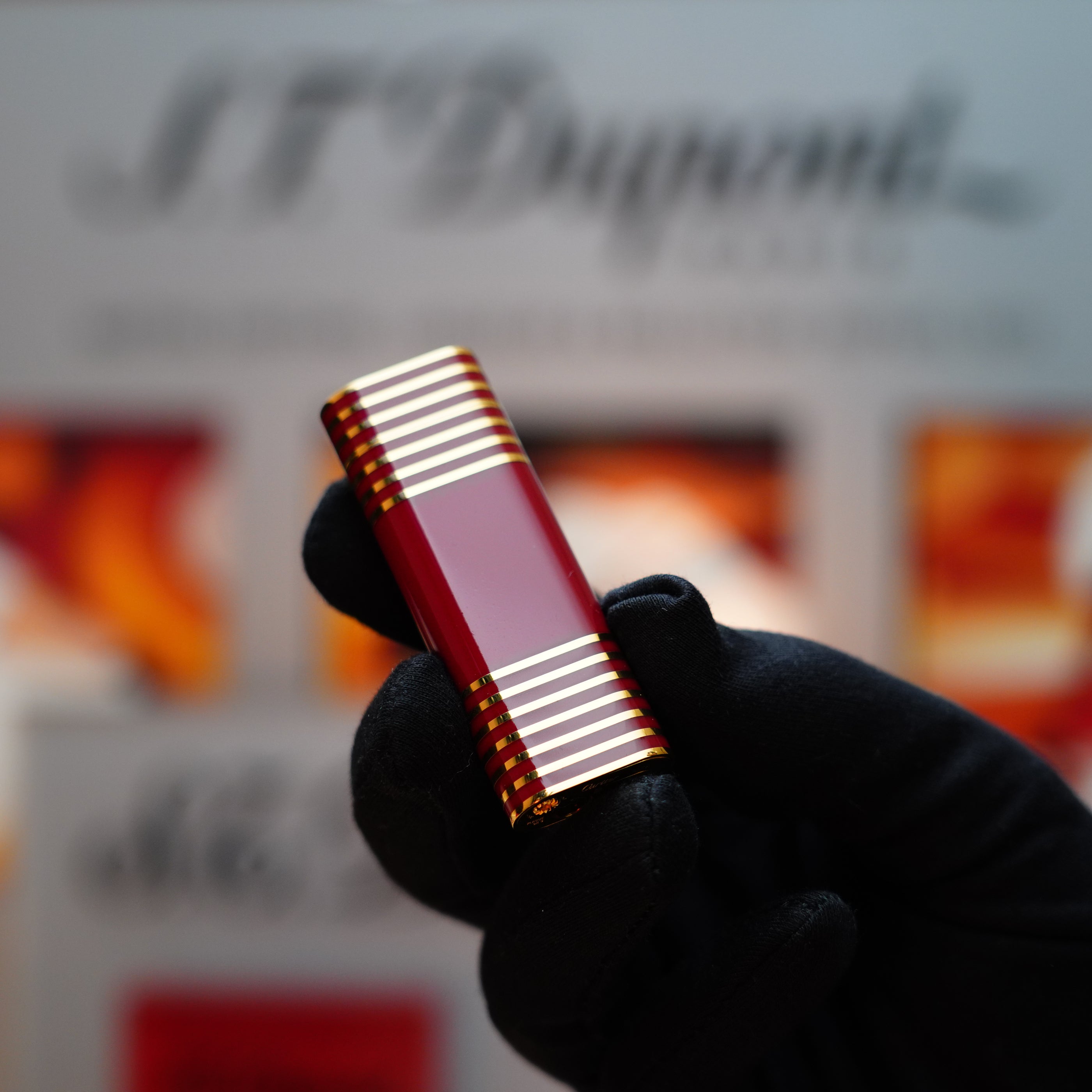 A gloved hand holds a Vintage 1985 Cartier Burgundy Red 24k Gold Lacquer Le Must Lighter, featuring a rare horizontal line pattern, gleaming against a blurred background.