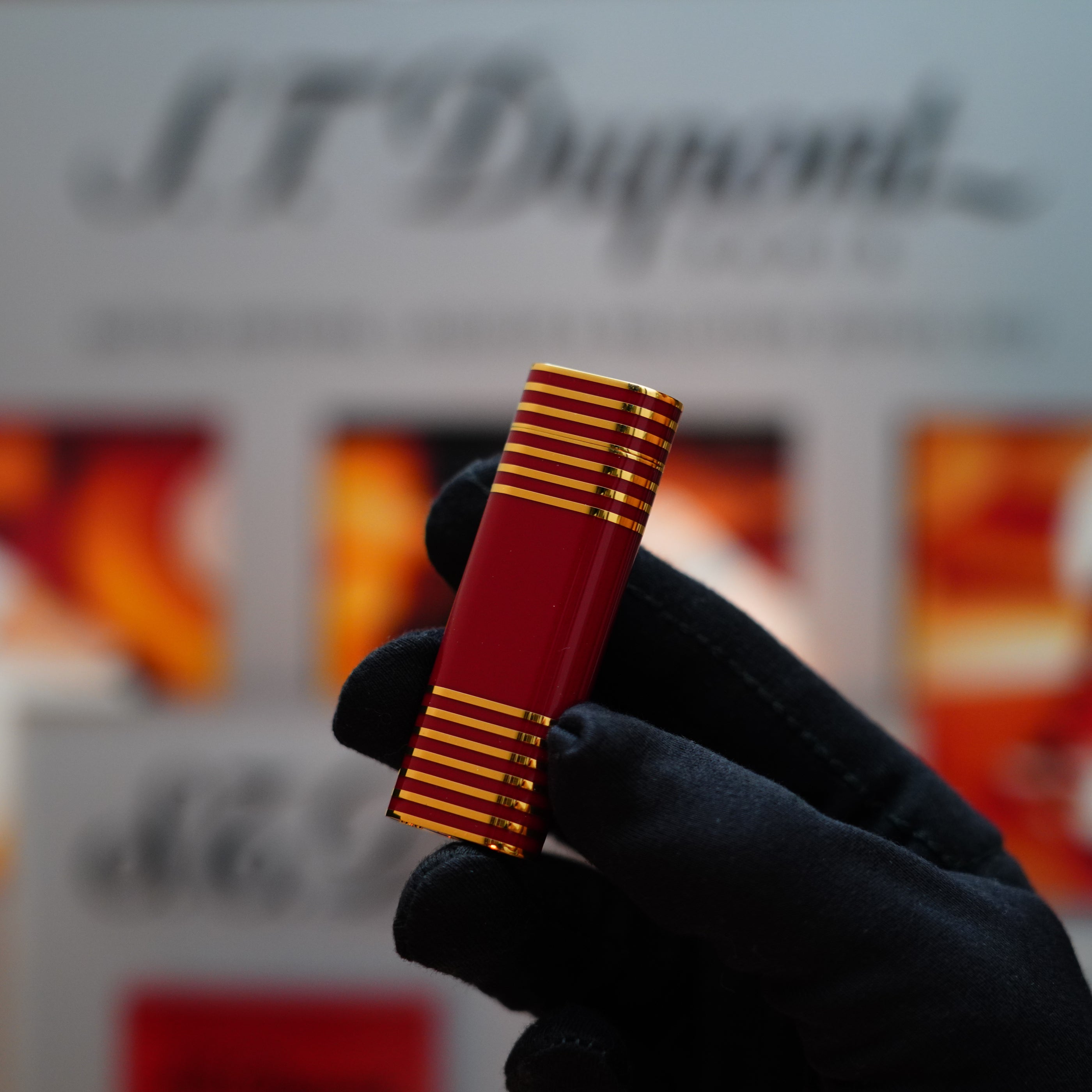 A gloved hand holds a Vintage 1985 Cartier Burgundy Red 24k Gold Lacquer Le Must Lighter adorned with a horizontal line pattern, set against a blurred background.