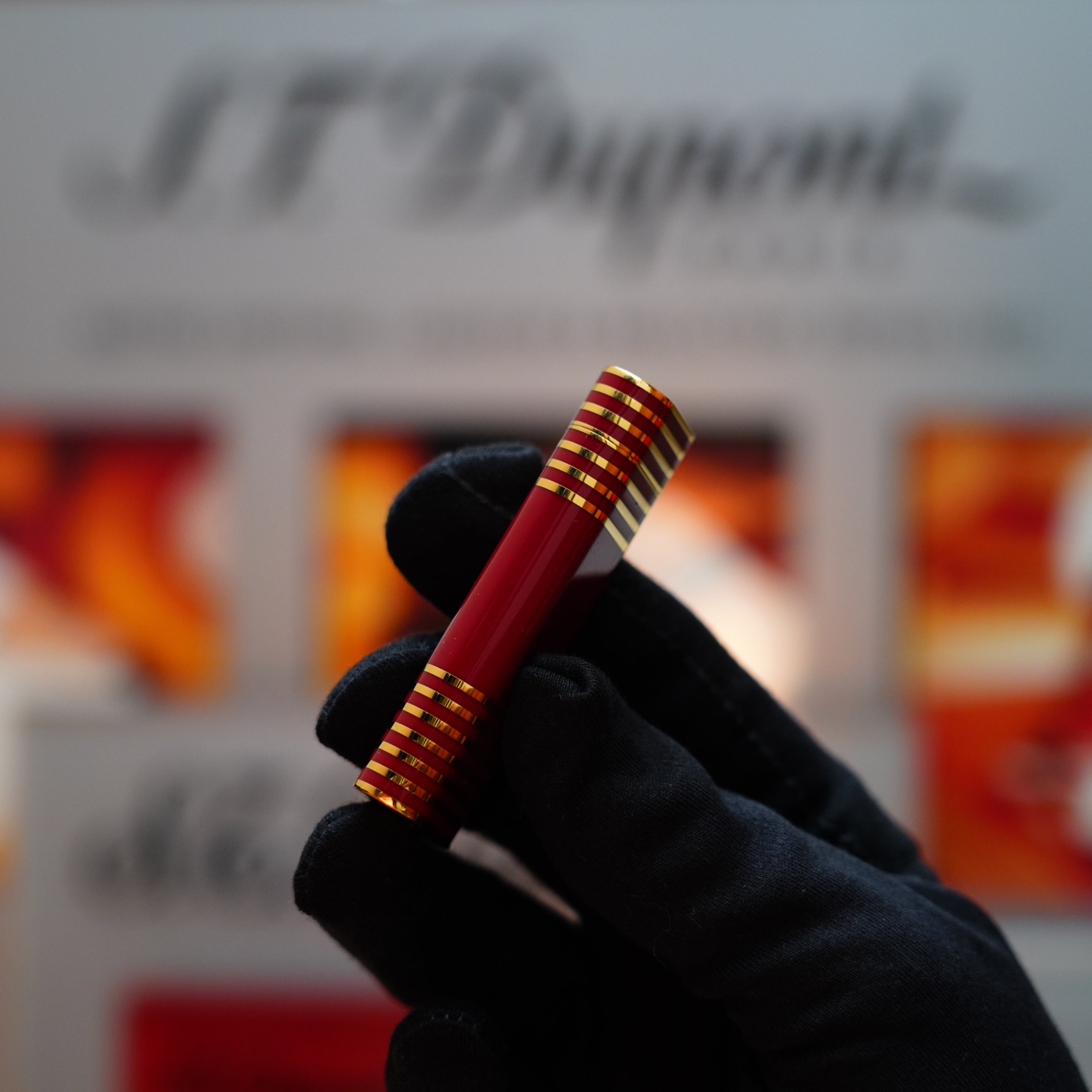 A gloved hand holds a burgundy red and gold cylindrical object, possibly a Vintage 1985 Cartier Burgundy Red 24k Gold Lacquer Le Must Lighter with the rare horizontal line pattern, against a blurred background. The text "Cartier" is visible, evoking the allure of fine craftsmanship akin to S.T. Dupont lighters.