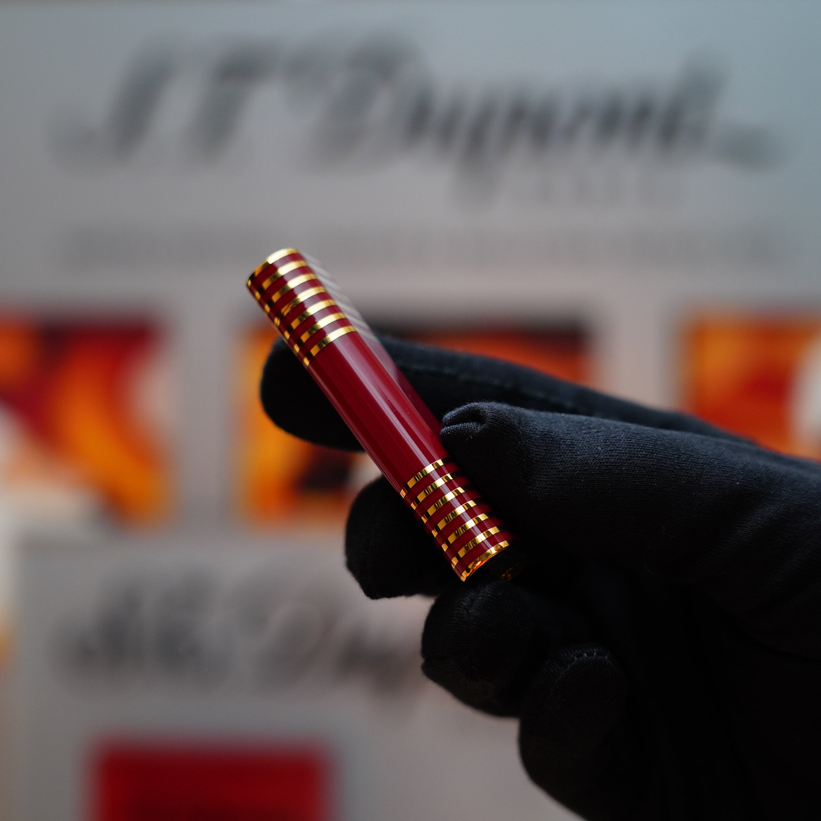 A gloved hand holding a Vintage 1985 Cartier Burgundy Red 24k Gold Lacquer with horizontal lines Le Must Lighter is set against a blurred background with text.