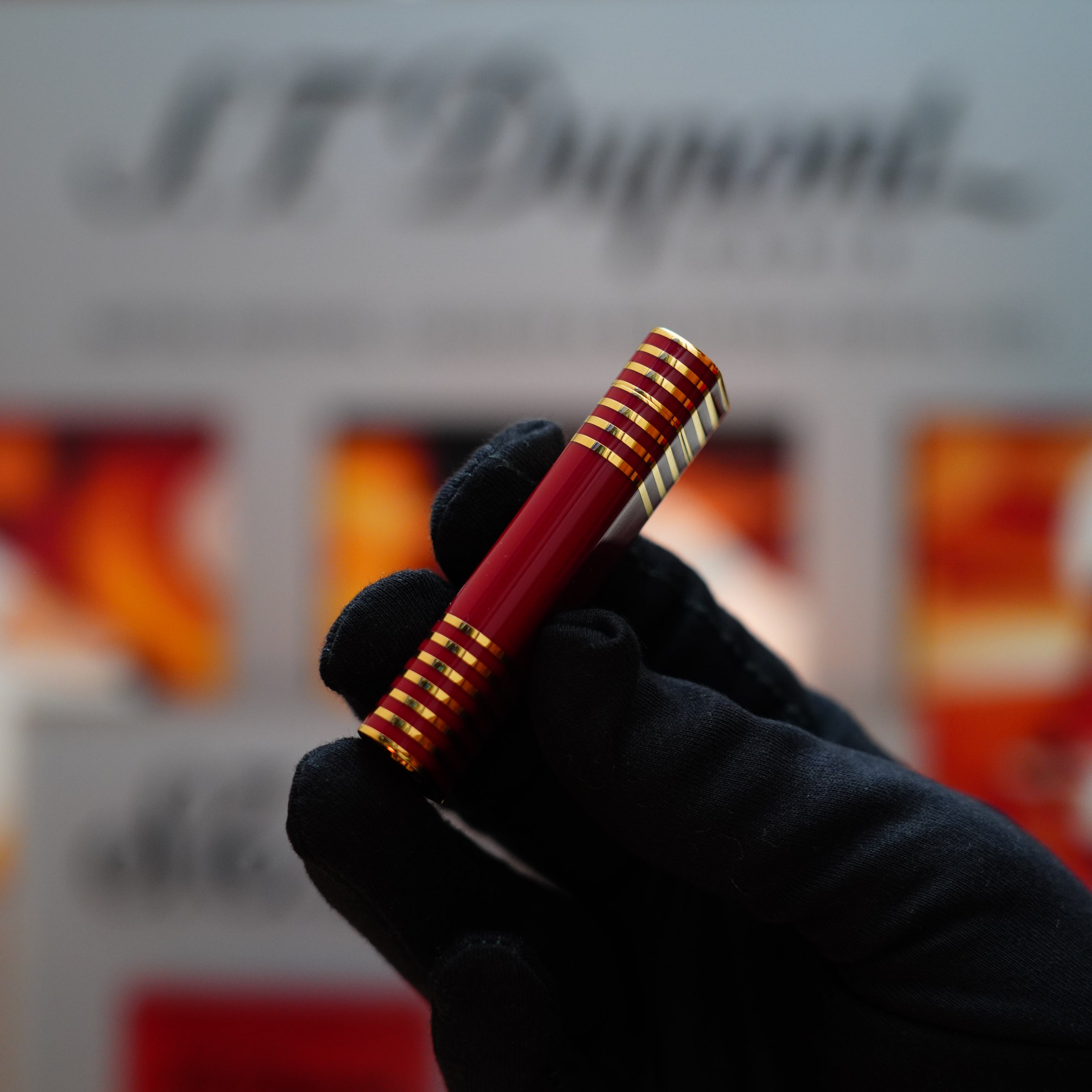 A gloved hand elegantly holds the Vintage 1985 Cartier Burgundy Red 24k Gold Lacquer Le Must Lighter, featuring horizontal lines, reminiscent of a vintage Cartier design, in front of a blurred background with text.