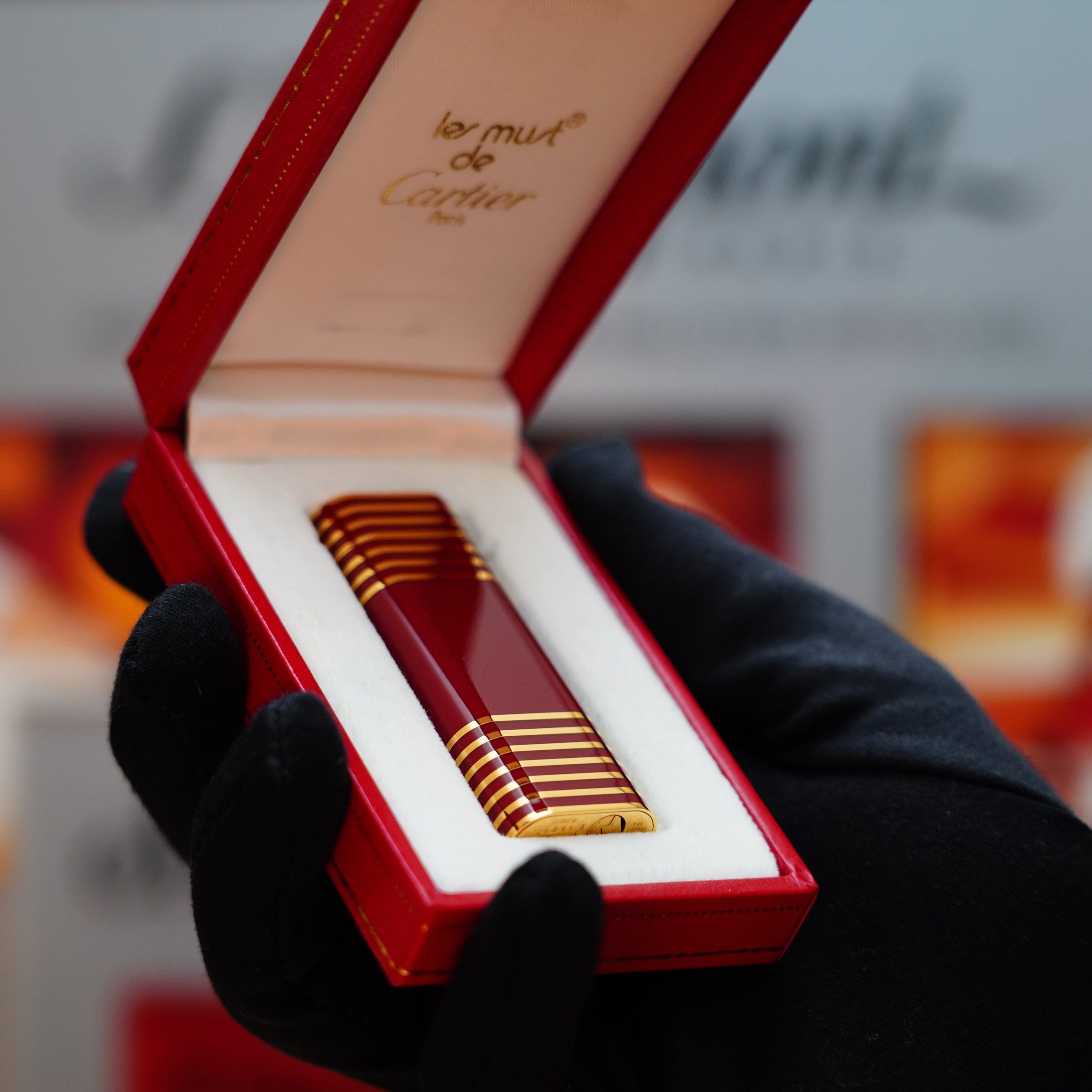 A gloved hand delicately holds a Vintage 1985 Cartier Burgundy Red Le Must Lighter, with a 24k gold lacquer and an exquisite horizontal line pattern, gracefully resting in its red box.