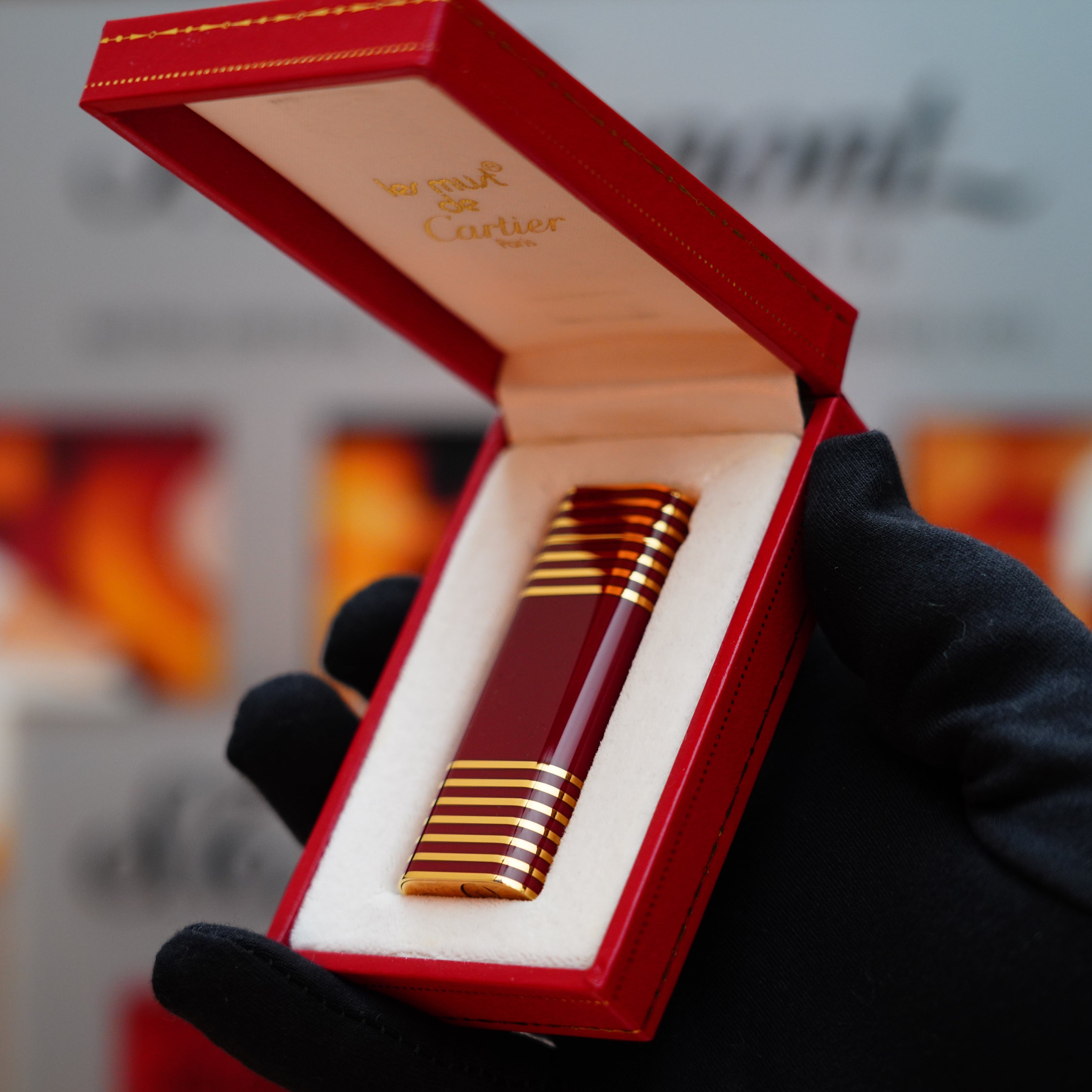 A gloved hand holds an open red Cartier box, revealing a sleek Vintage 1985 Cartier Burgundy Red 24k Gold Lacquer Le Must Lighter, adorned with a rare horizontal line pattern and elegant gold accents.