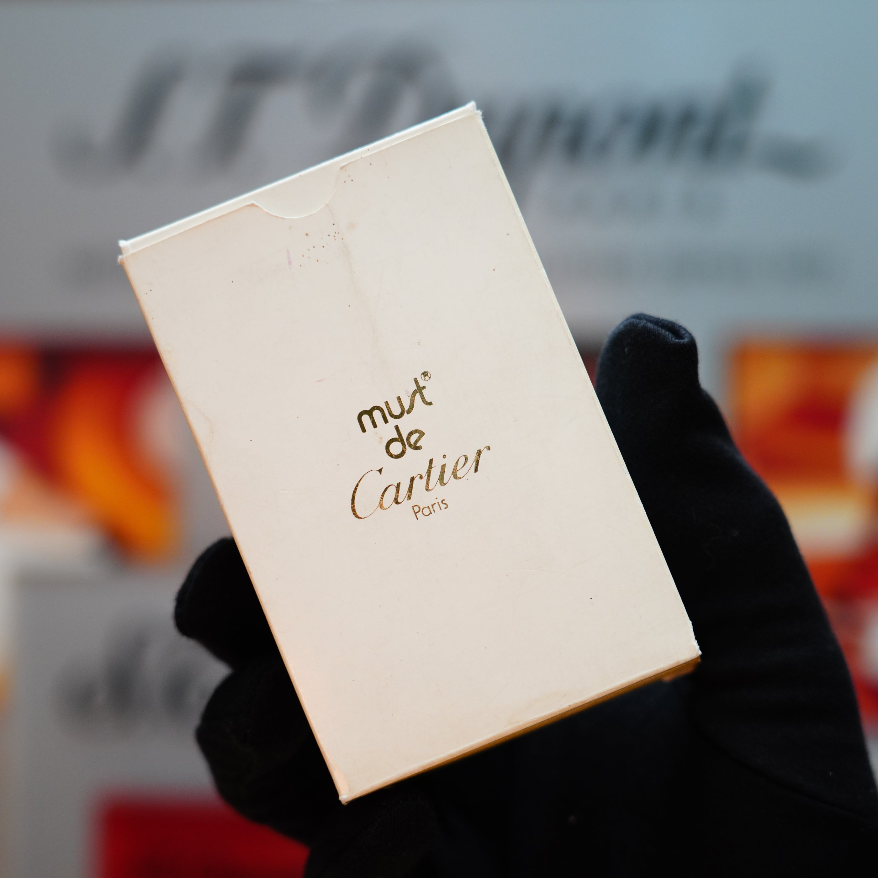 A gloved hand holds a white box labeled "Must de Cartier Paris" against a blurred background, suggesting the allure of a rare Vintage 1985 Cartier Burgundy Red 24k Gold Lacquer Le Must Lighter adorned with its iconic horizontal line design.