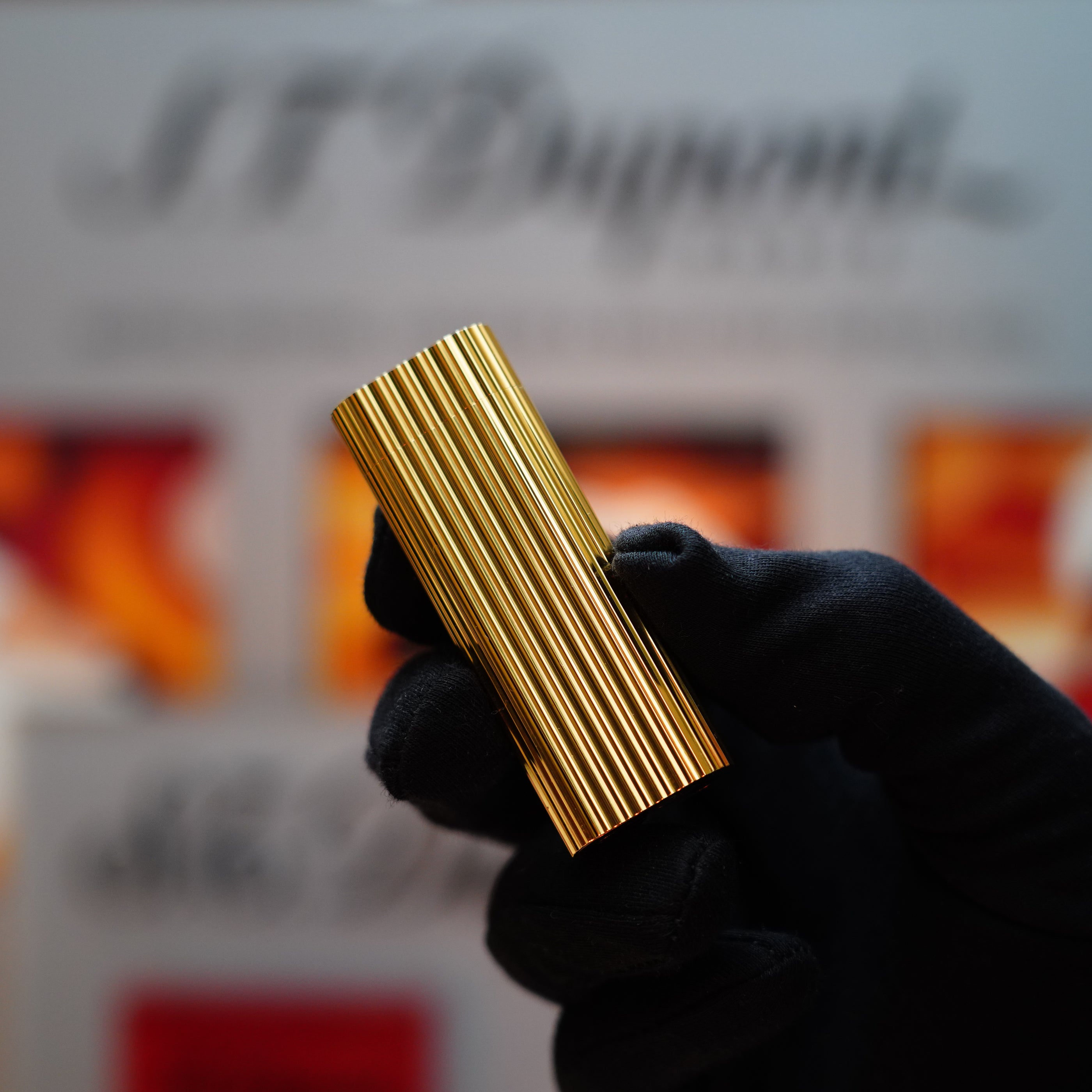 A gloved hand holds a vintage 1980 Cartier 24k Gold Finish Double Vertical Line Le Must Lighter against a blurred background with text, capturing the elegance of a Cartier lighter.