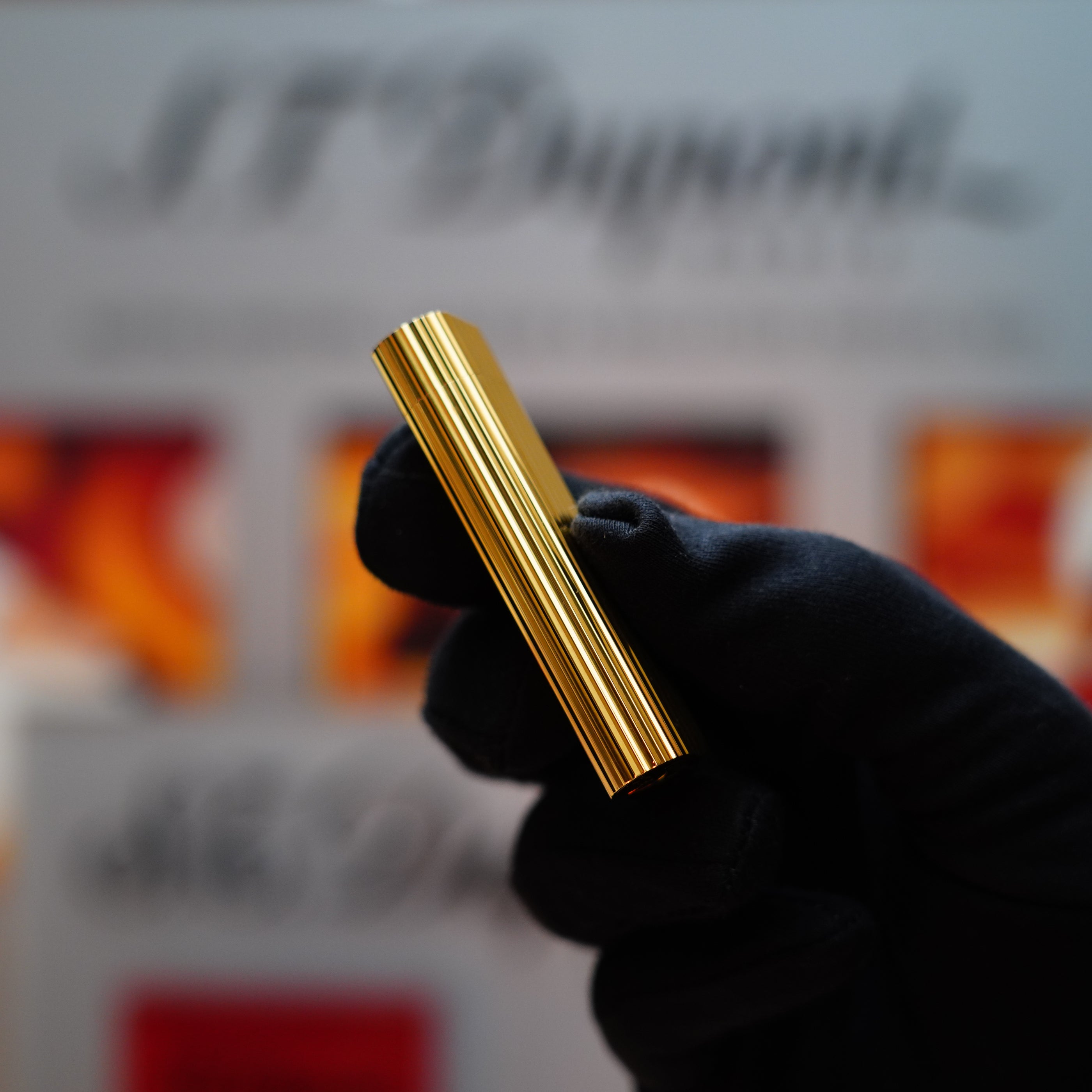 A gloved hand holds a sleek Vintage 1980 Cartier 24k Gold Finish Double Vertical Line Le Must Lighter in focus, with a blurred background of signage and colors.