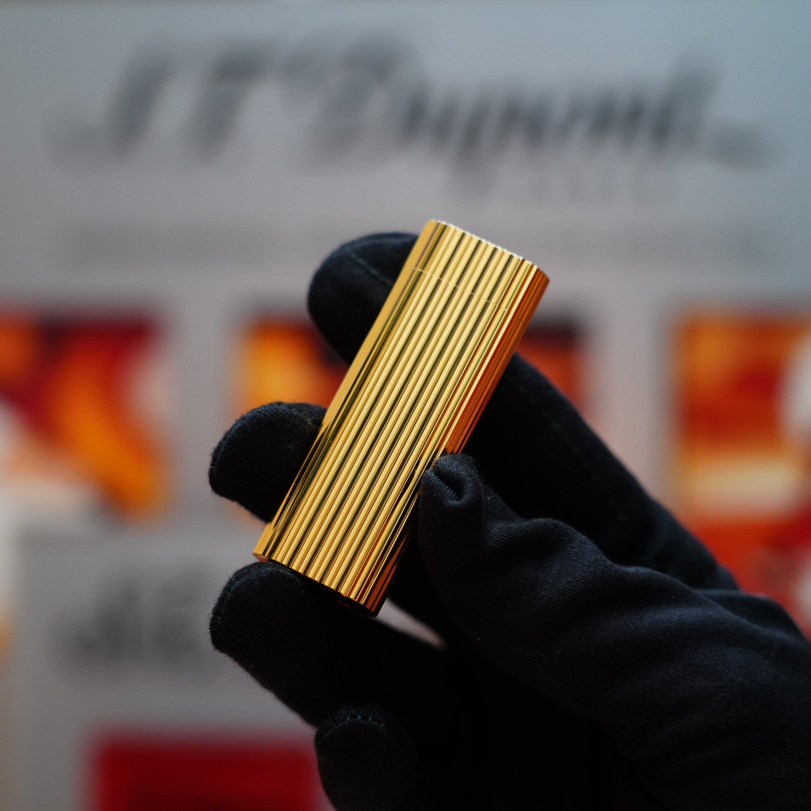 A gloved hand holds a rectangular object with gold stripes, likely the Vintage 1980 Cartier 24k Gold Finish Double Vertical Line Le Must Lighter, against a blurred background featuring the Cartier logo.