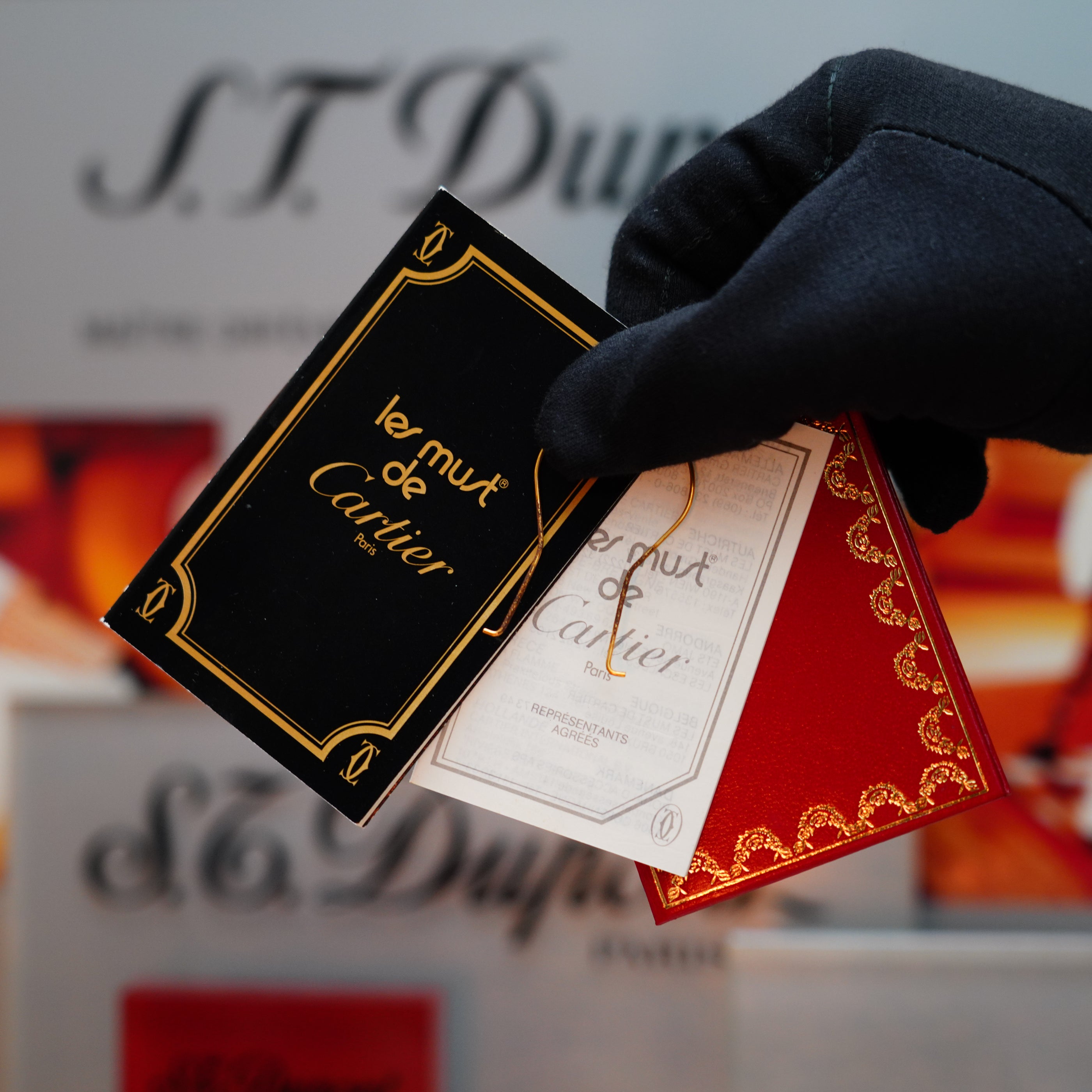 A gloved hand displays three perfume cards labeled "le must de Cartier" in black, white, and red. The blurred background subtly reveals a hint of "S.T. Dupont" signage, evoking the allure of a collector's item such as the Vintage 1980 Cartier 24k Gold Finish Double Vertical Line Le Must Lighter by Cartier.