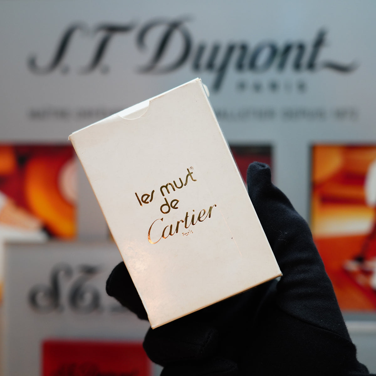 A gloved hand holding a small white box labeled "le must de Cartier" set against a blurred background showcasing S.T. Dupont branding captures the charm of the Vintage 1980 Cartier 24k Gold Finish Double Vertical Line Le Must Lighter—a true collector's piece.