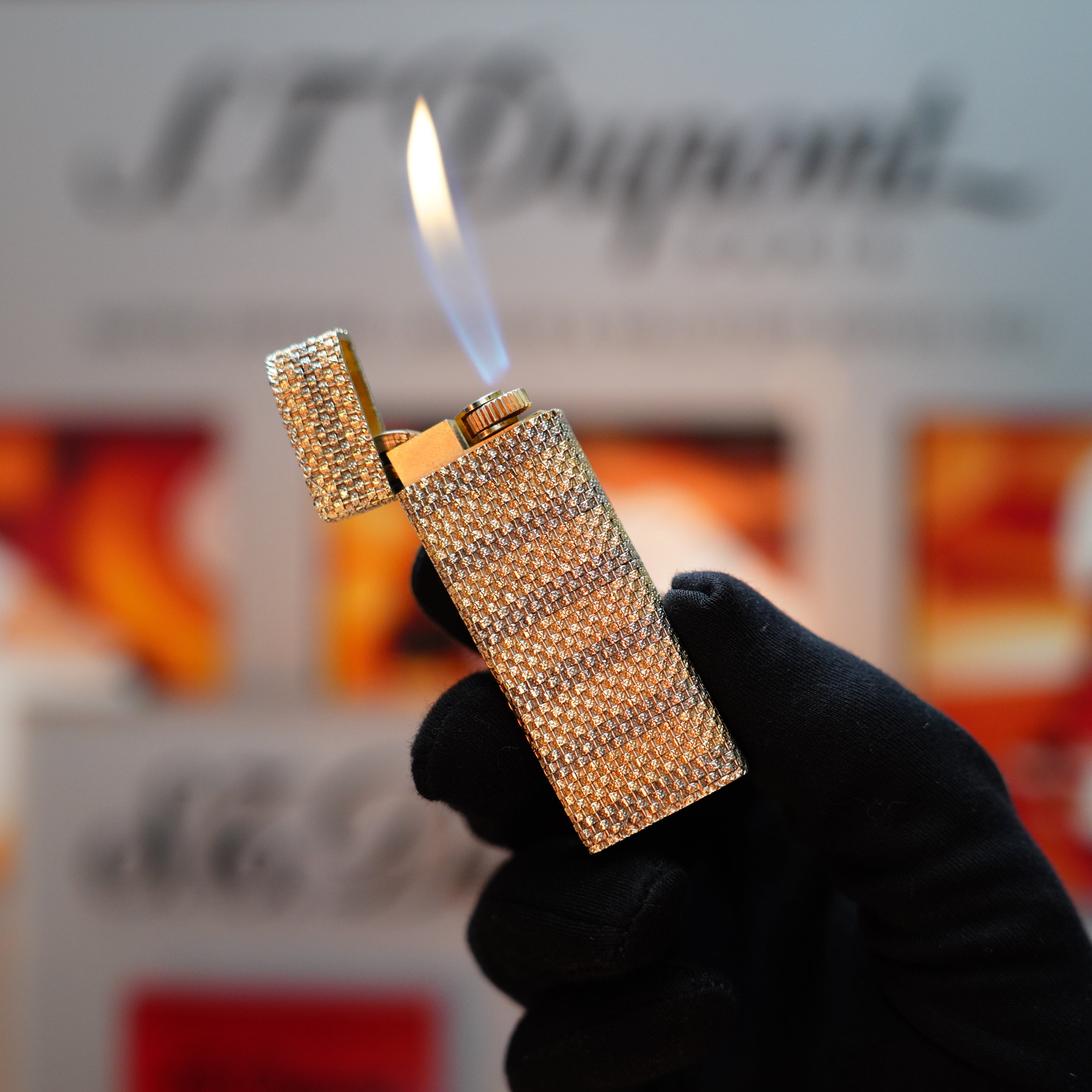 A gloved hand delicately holds an ornate Vintage 1978 Cartier Rare Pentagon Solid 18k Gold Jacket Basket with a lit flame, set against a blurred background of text and colorful images. This exquisite piece from Cartier, adorned with 750 hallmarks, exudes timeless elegance and craftsmanship.