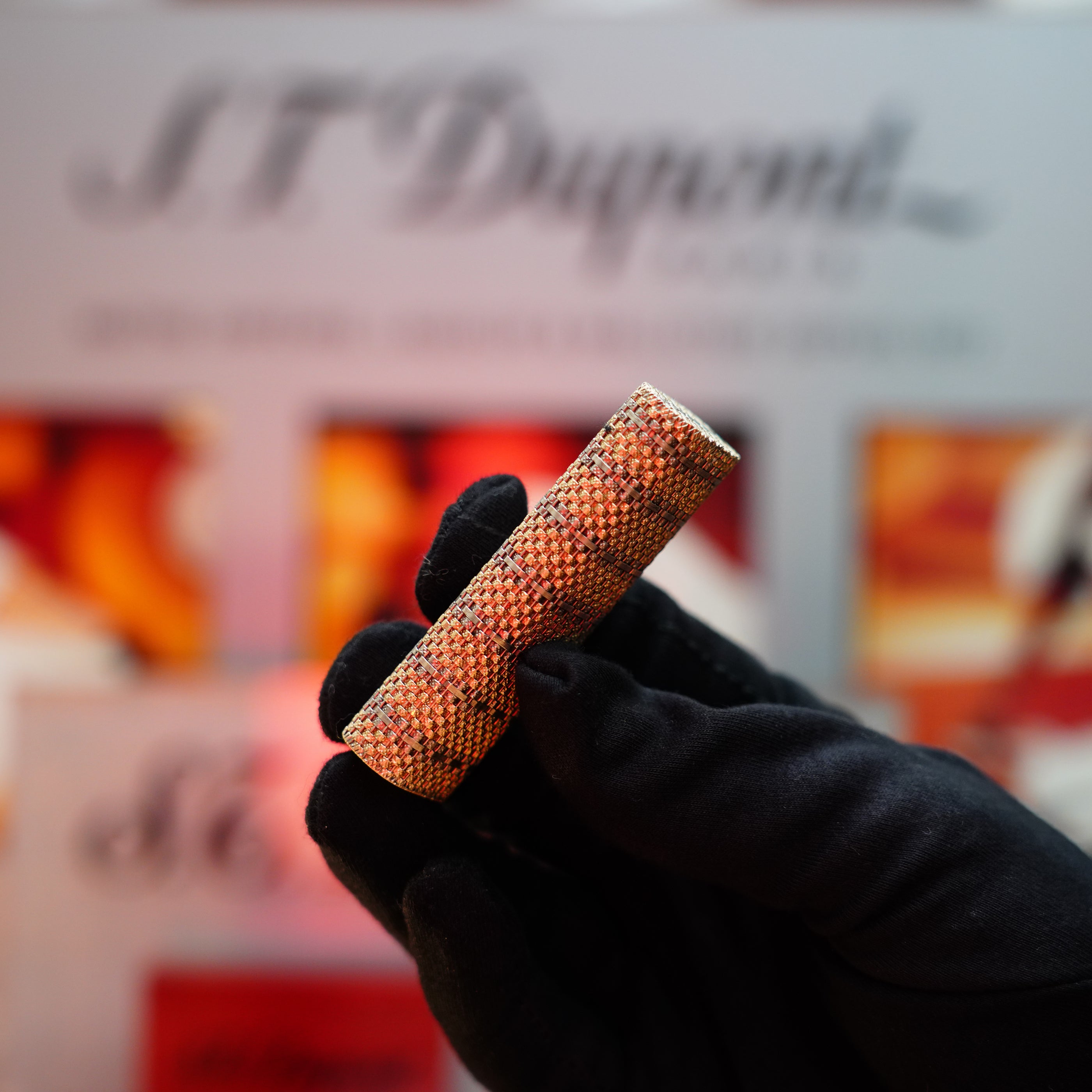 A gloved hand holding the Vintage 1982 Cartier Solid 18k Gold Jacket Basket Lighter, featuring a textured surface similar to a classic Cartier design. The background is blurred, with indistinct text and colors.