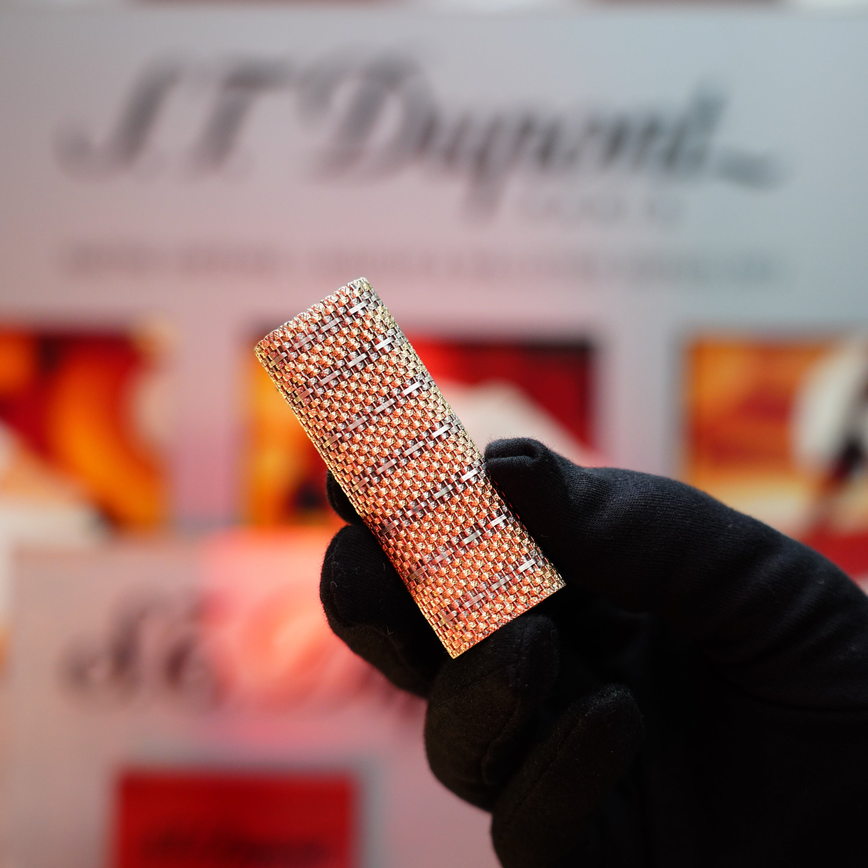 A gloved hand holds a rectangular, textured object, potentially a Vintage 1982 Cartier Solid 18k Gold Jacket Basket Lighter with 750 hallmarks, in front of a blurred background featuring indistinct lettering and images.