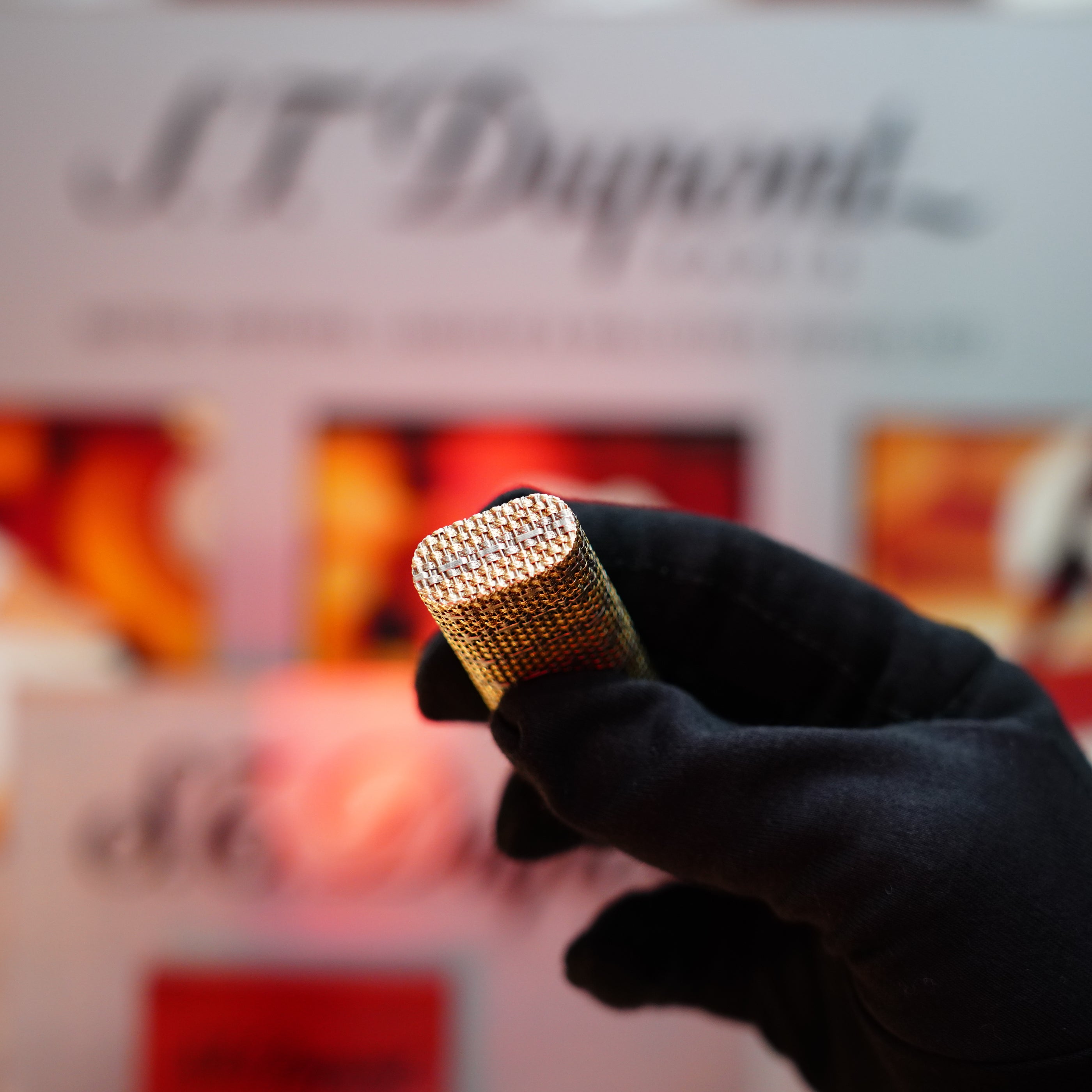 A person wearing black gloves holds a rare collectible—a cylinder made of solid 18k gold, known as the Vintage 1982 Cartier Jacket Basket Lighter with 750 hallmarks—in front of a blurred background with the text "Cartier.