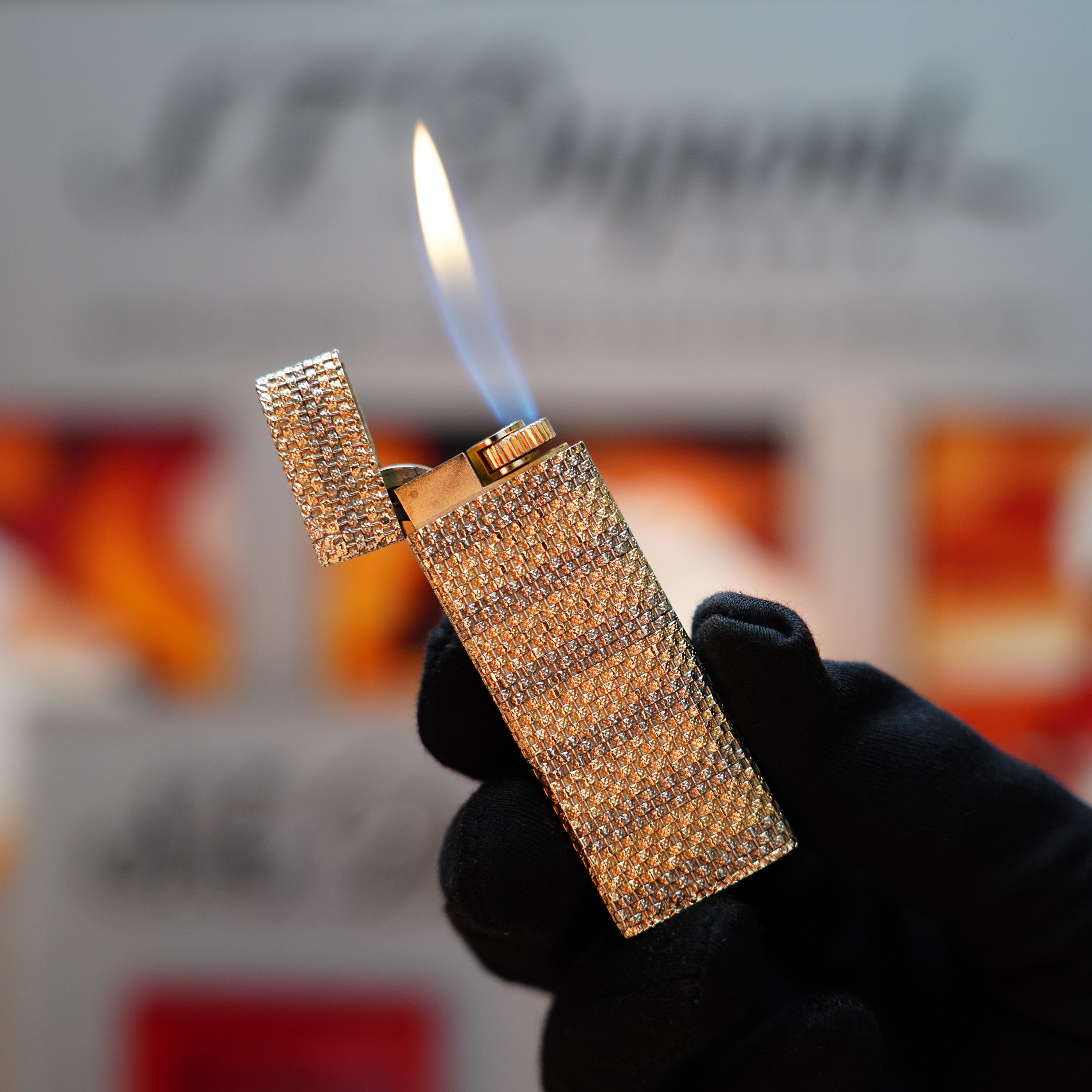 A hand wearing a black glove holds an open, vintage Cartier lighter from 1978's Rare Pentagon series, shimmering in solid 18k gold with a diamond-encrusted design and a vibrant blue flame.