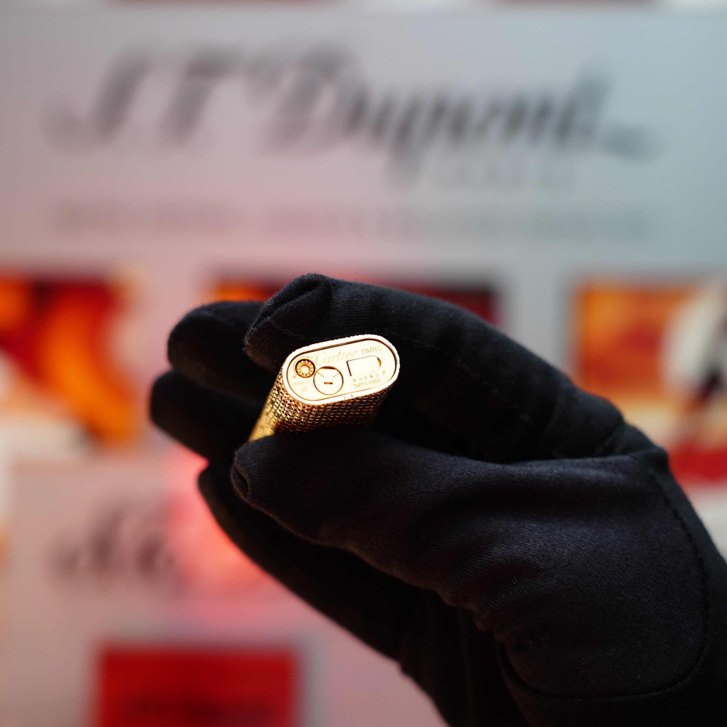 A gloved hand holds a rare collectible gold metallic lighter, the "Cartier" brand subtly gleaming in the background, reminiscent of the elegance of a Vintage 1982 Cartier Solid 18k Gold Jacket Basket Lighter with 750 hallmarks.