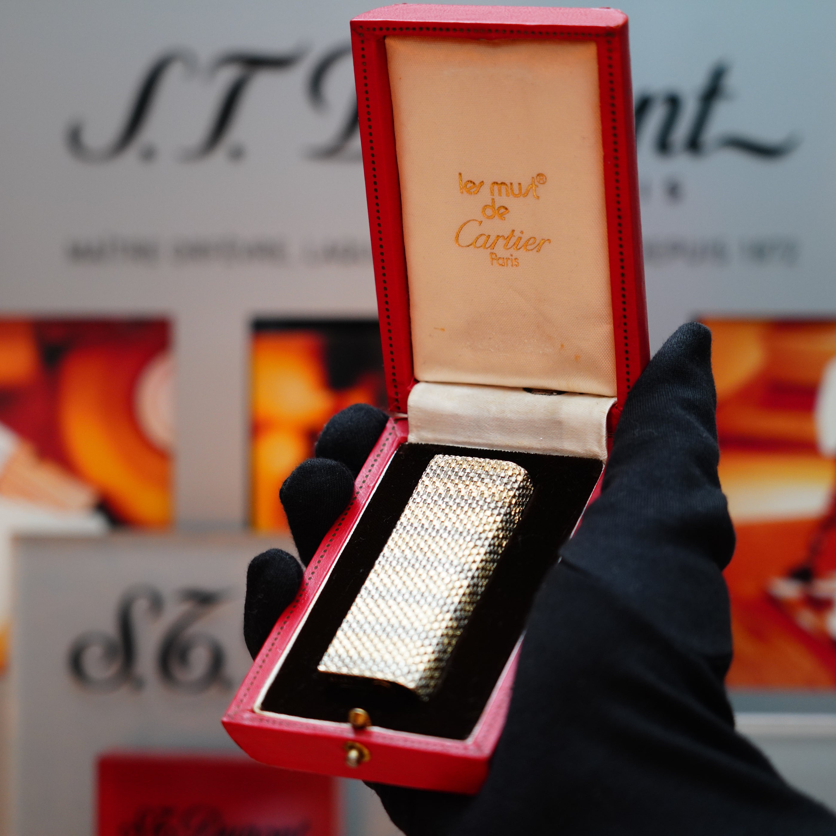 A gloved hand holds a vintage Cartier Pentagon lighter from the rare Jacket Basket series, crafted from solid 18k gold and marked with 750 hallmarks, nestled in a red Cartier box, with a blurred background displaying text and images.