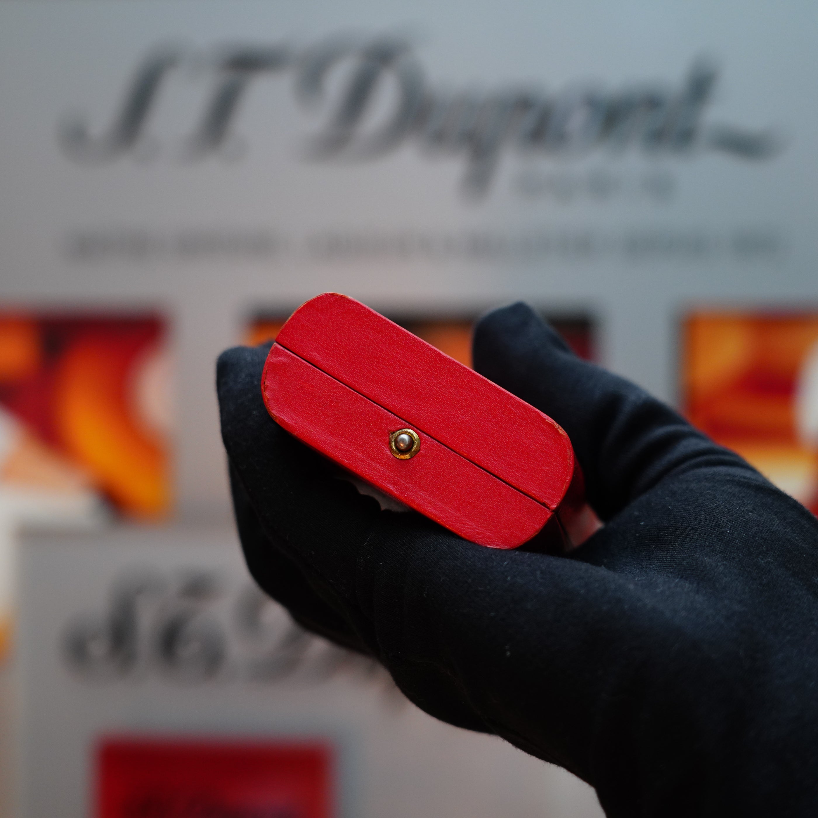 A gloved hand holds a striking red object, reminiscent of the Vintage 1978 Cartier Rare Pentagon Solid 18k Gold Jacket Basket Lighter with 750 hallmarks, in front of a blurred S.T. Dupont logo background.