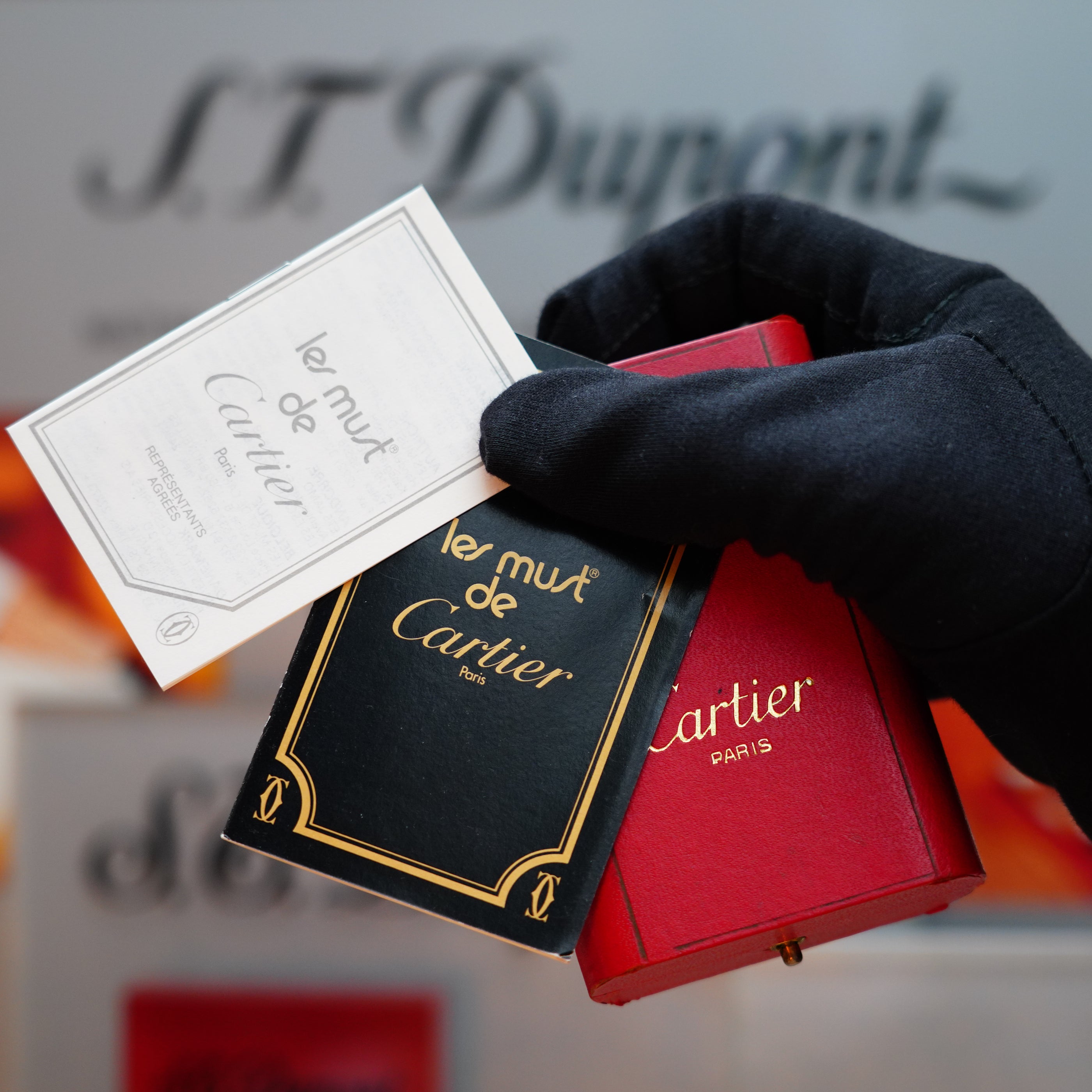 A gloved hand elegantly holds a black and red Cartier box, adorned with the label "le must de Cartier." Inside, you'll find the Vintage 1978 Cartier Rare Pentagon Solid 18k Gold Jacket Basket Lighter with 750 hallmarks.