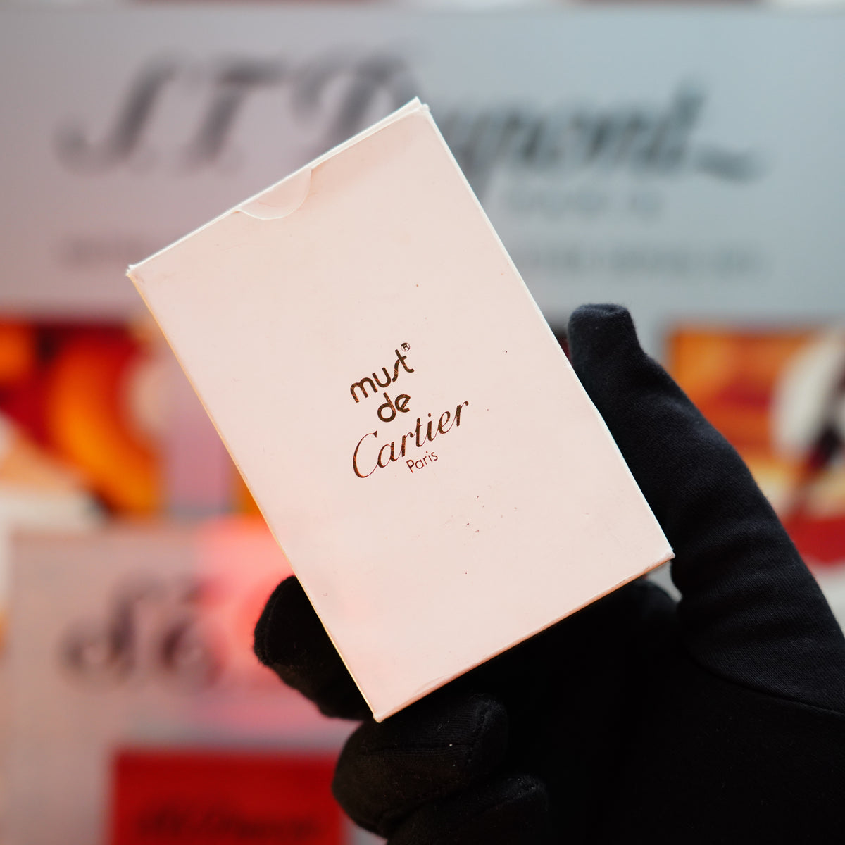 A gloved hand holds a white box adorned with "Must de Cartier Paris," suggesting the exquisite charm of the Vintage 1982 Cartier Solid 18k Gold Jacket Basket Lighter—a rare collectible from the renowned brand, distinguished by its luxurious solid gold design featuring 750 hallmarks, all set against a softly blurred background.